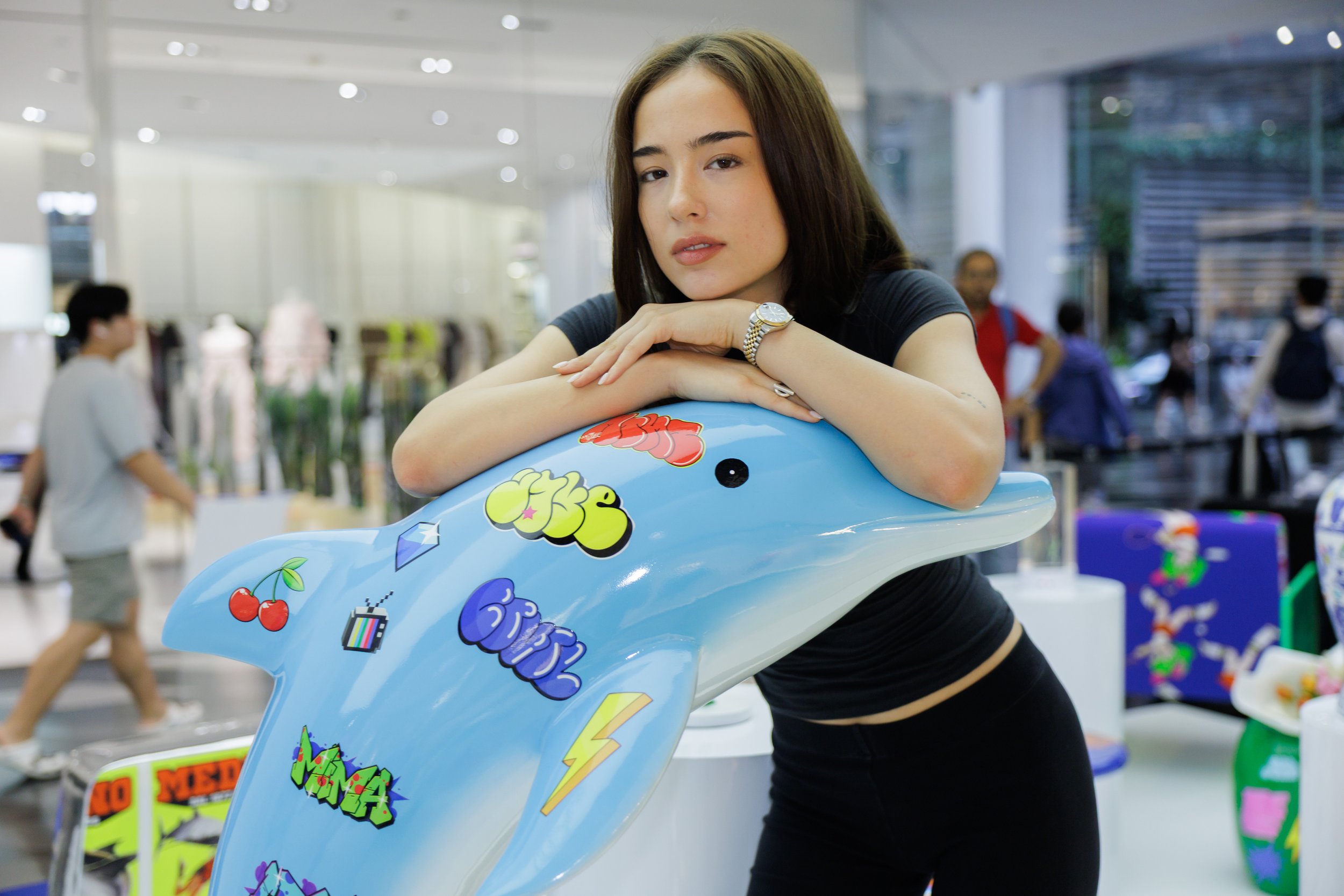 Young woman resting her chin on her arms while leaning on a inflatable dolphin at an indoor event or store with other people and display booths in the background.