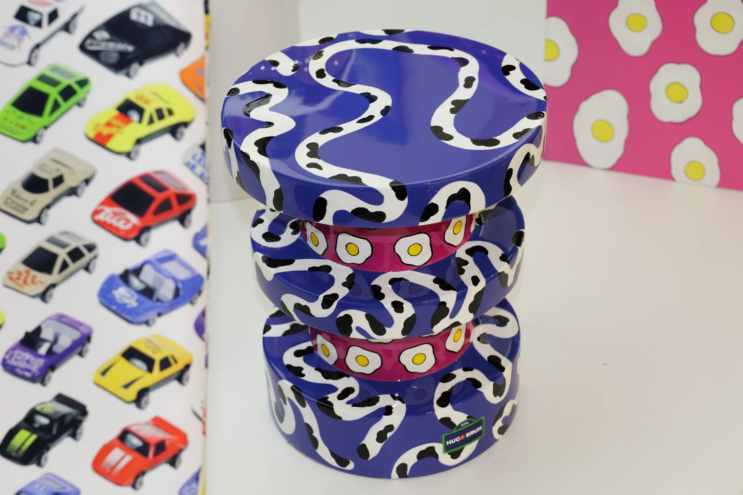 Stack of three colorful ceramic bowls with a leopard print pattern, placed on a white surface, with a background featuring a poster of toy cars and a pink decorative panel with fried egg patterns.