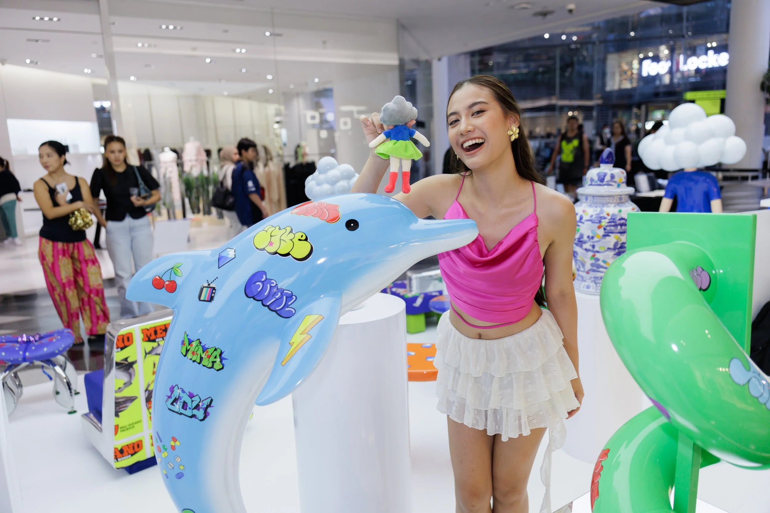 A young woman in a pink top and beige skirt is smiling and holding a small plush doll in front of colorful whale-themed displays at a shopping mall.