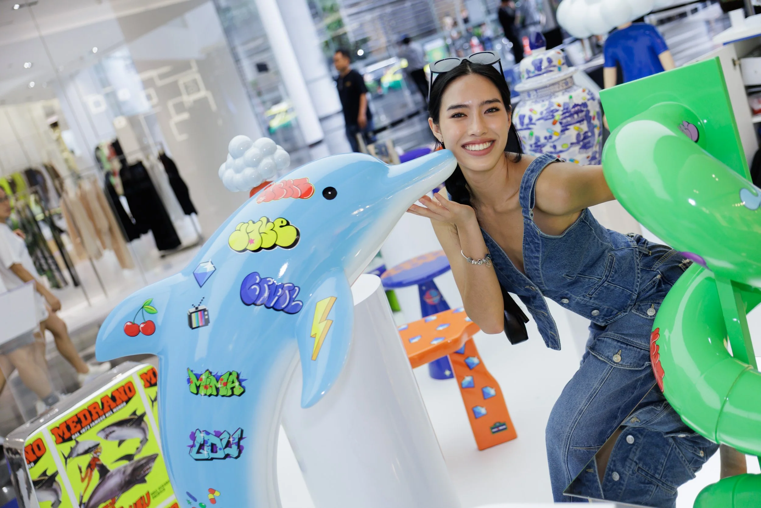 A woman smiling at an arcade surrounded by colorful arcade games and decorations. She has sunglasses on her head, wears a denim outfit, and is posing with a blue and green arcade game machine.