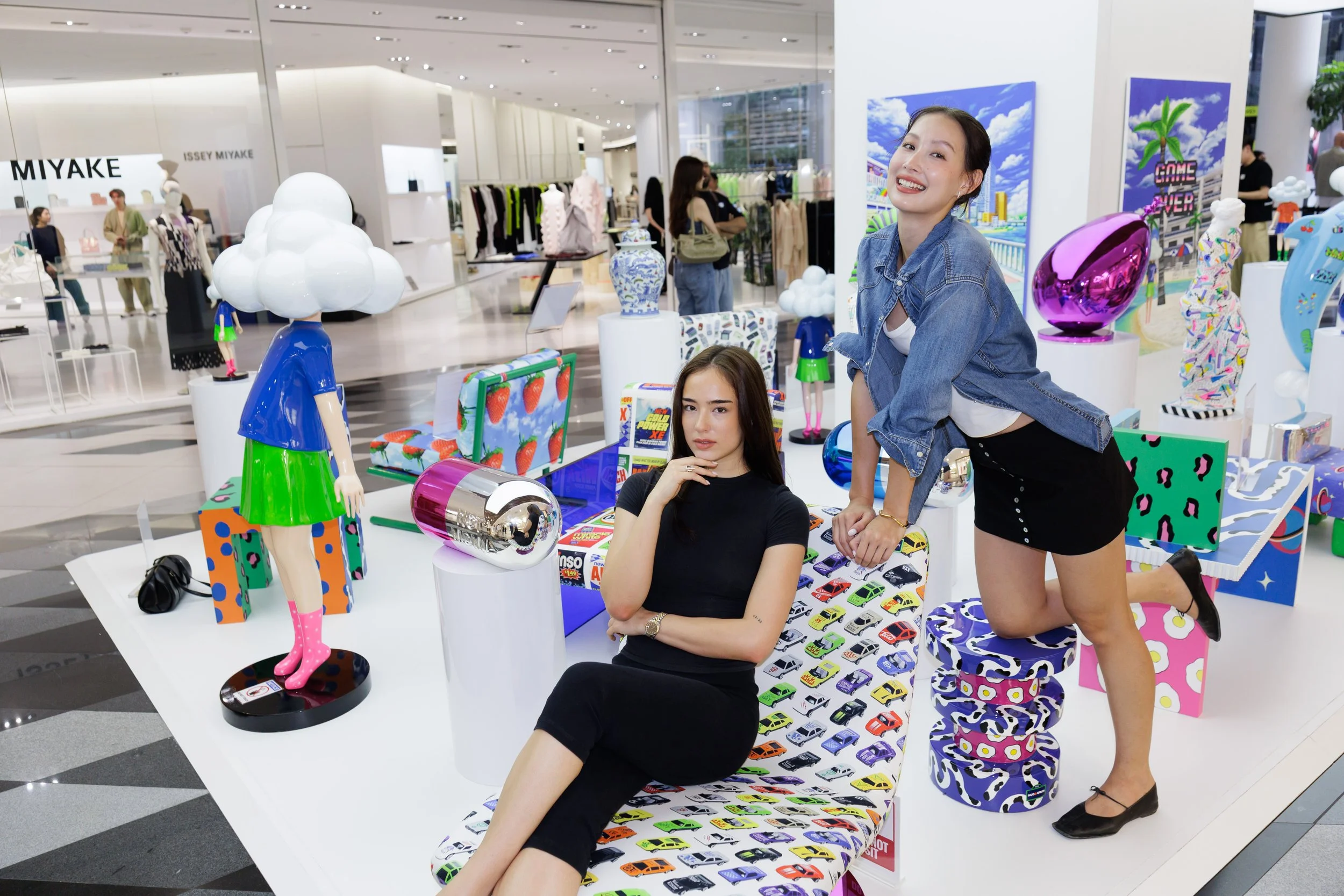Two women are sitting and kneeling at a colorful display table with various art sculptures and objects, inside a shopping mall featuring clothing stores and art displays in the background.