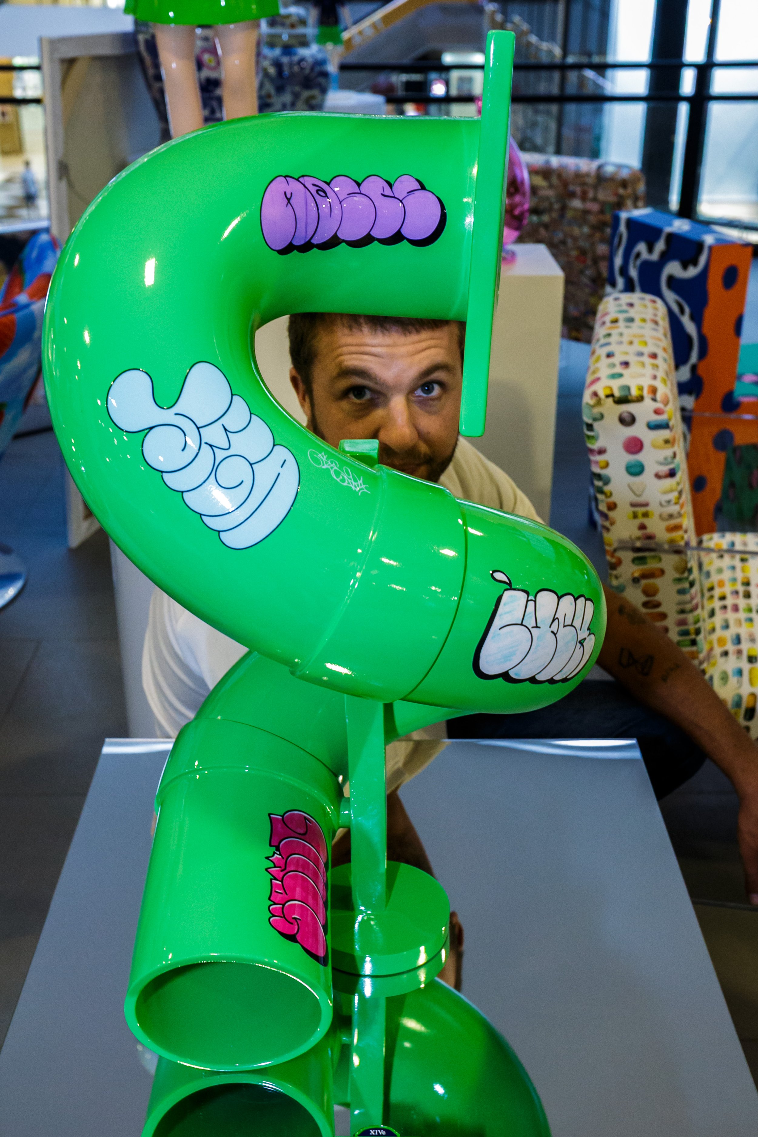 A man peeks through a bright green, graffiti-styled sculpture with bubble letters, in a store with colorful products in the background.