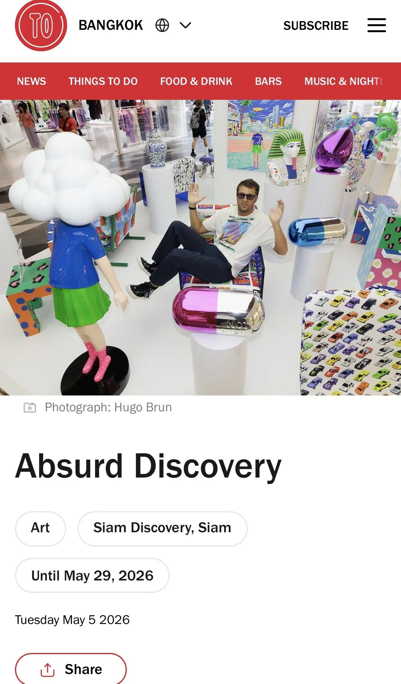 A man wearing sunglasses and a white T-shirt is sitting on a colorful chair in an art installation. The installation features a mannequin with a cloud-shaped head, wearing a blue shirt, green skirt, and pink boots, surrounded by various colorful sculptures and paintings, including a cityscape and abstract figures.