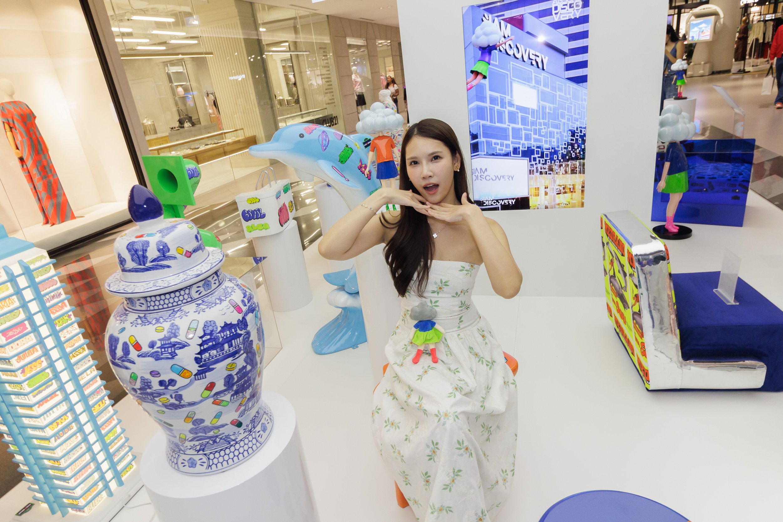 A young woman with long dark hair in a floral dress posing in front of colorful store displays. The background shows mannequins, a large digital screen, and shopping mall elements.