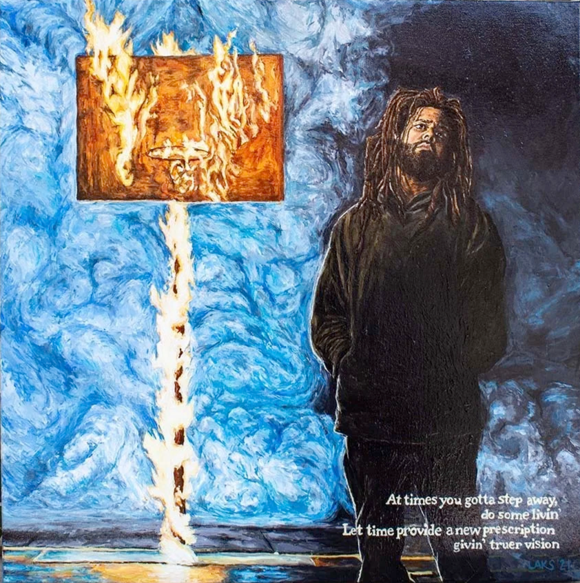 A man with dreadlocks and a beard, wearing a black hoodie, stands with his hands in his pockets, in front of a blue textured background. To his left is a framed picture of a face on fire, with flames extending downward.