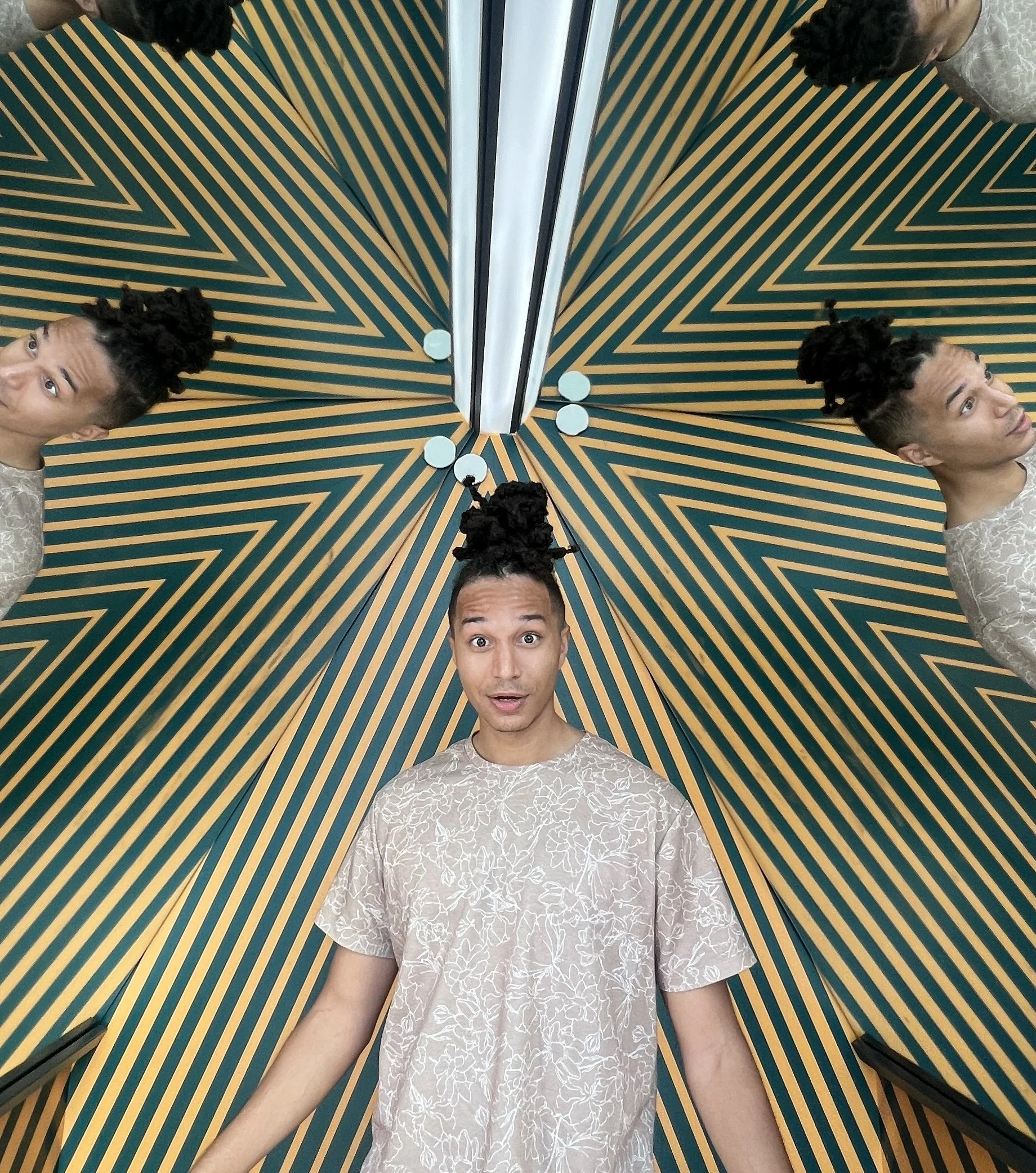 A man standing in front of a mirror, taking a selfie, with the mirror creating a symmetrical reflection. The background has a geometric pattern with yellow and green diagonal lines. The man has styled hair and is wearing a light-colored shirt with a subtle floral pattern.
