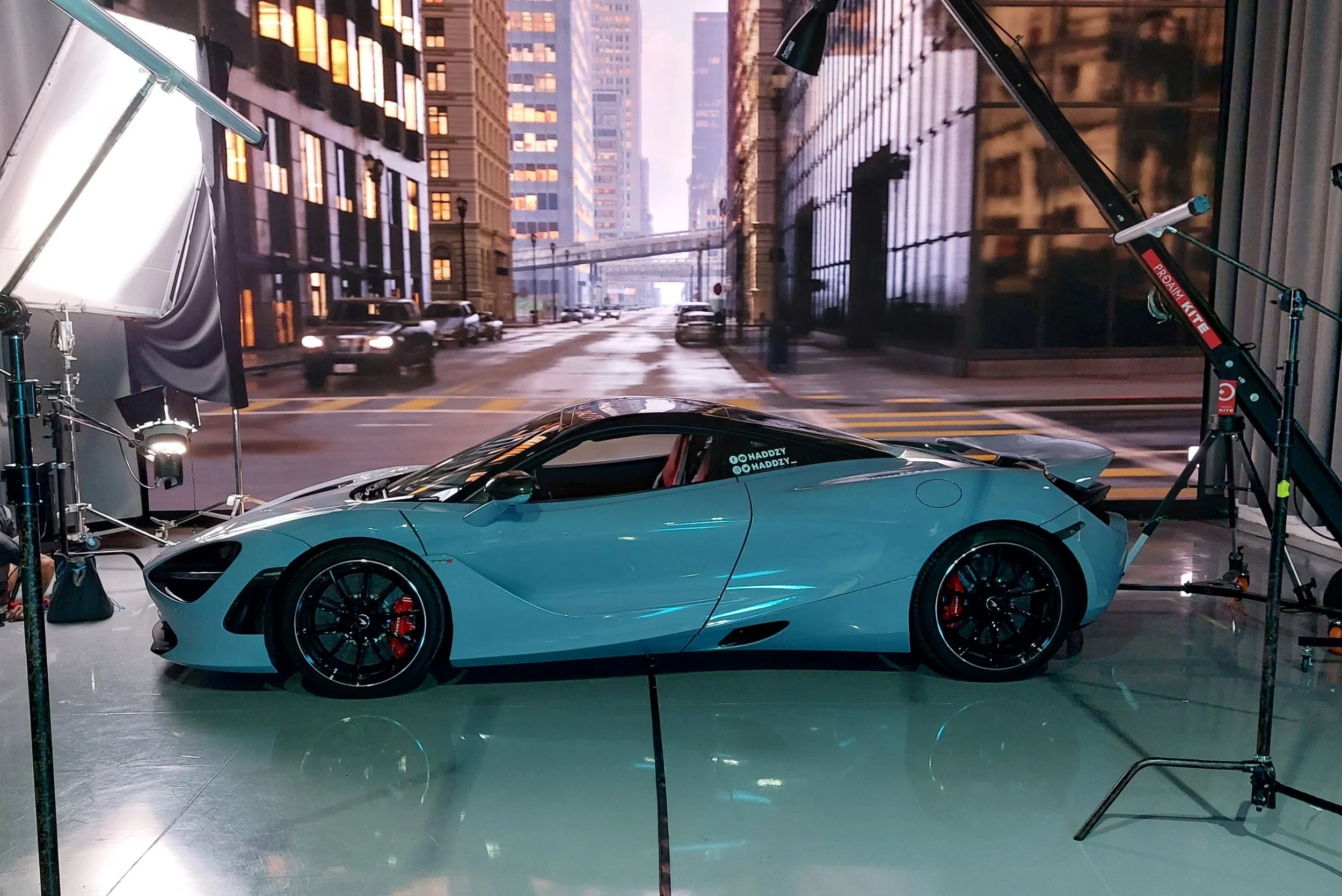 A blue sports car is inside a photography studio with city view backdrop, lighting equipment, and support stands.