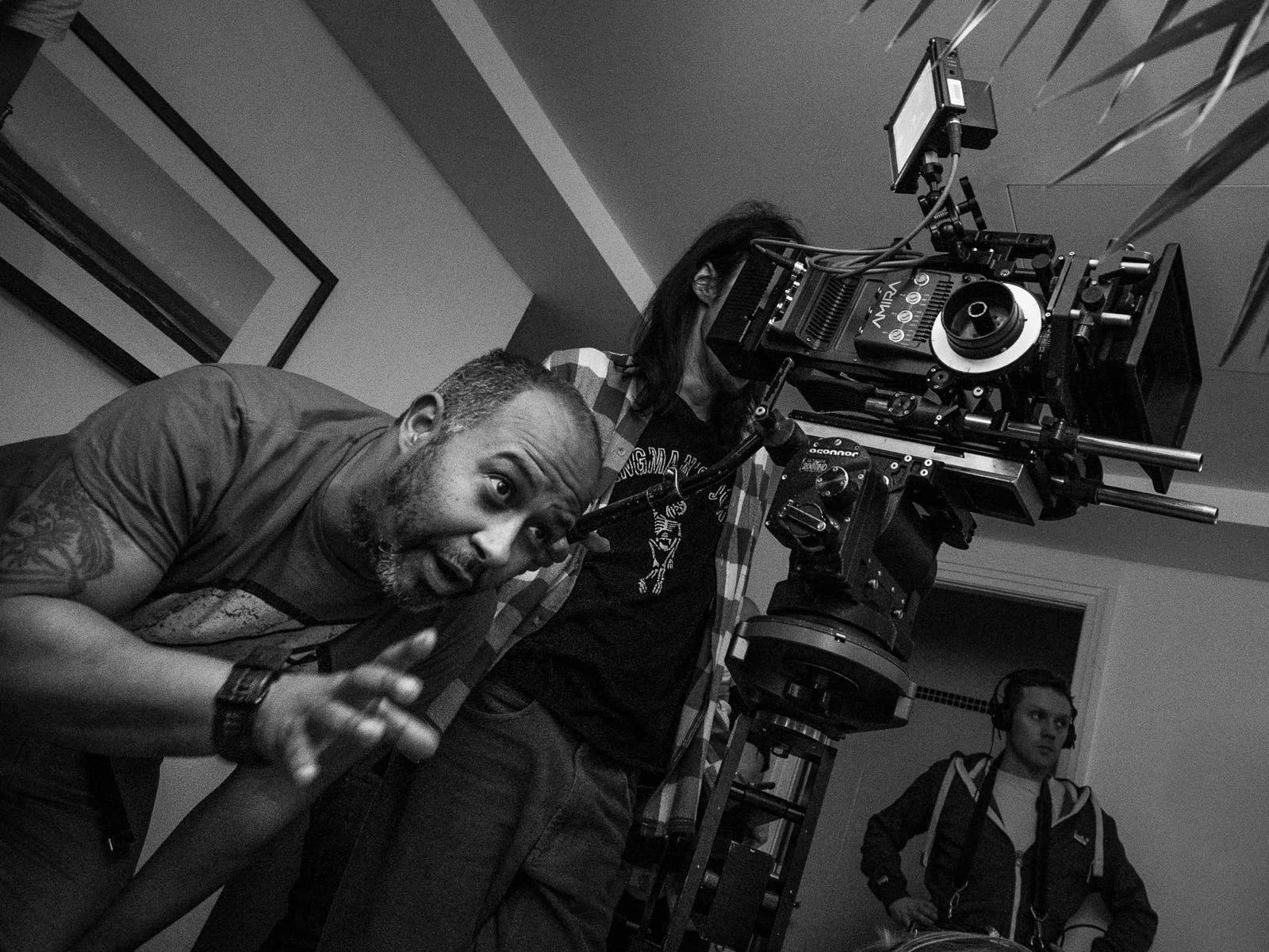 Black and white photo of three people behind a professional film camera. One person is leaning forward and pointing, appearing to give direction. Another is adjusting the camera, and a third stands observing, all indoors.