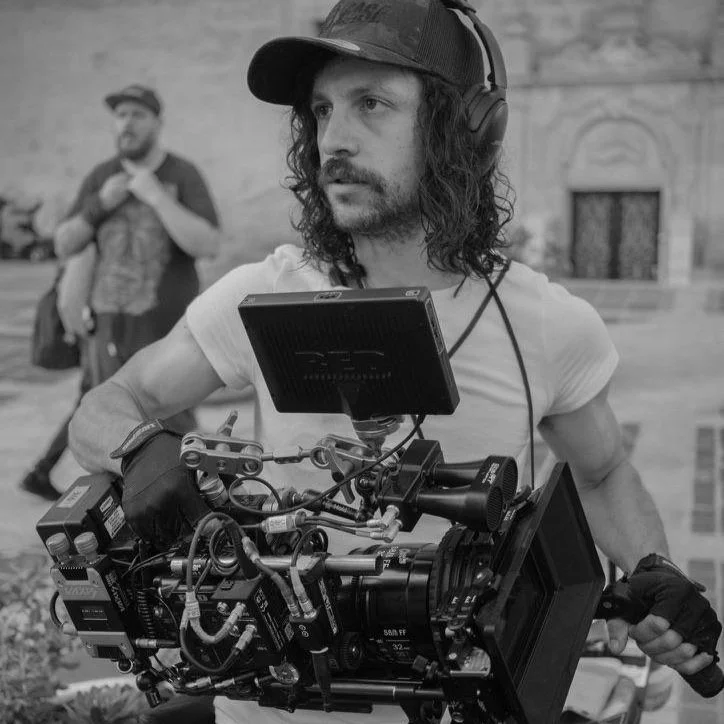 A man with long curly hair wearing a cap, headphones, and a white t-shirt holding a professional camera setup with a monitor, while another person stands in the background.