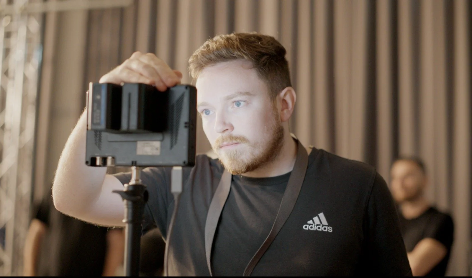 A man with a beard and blue eyes taking a photo or video with a camera on a tripod, in an indoor setting with gray curtains in the background.