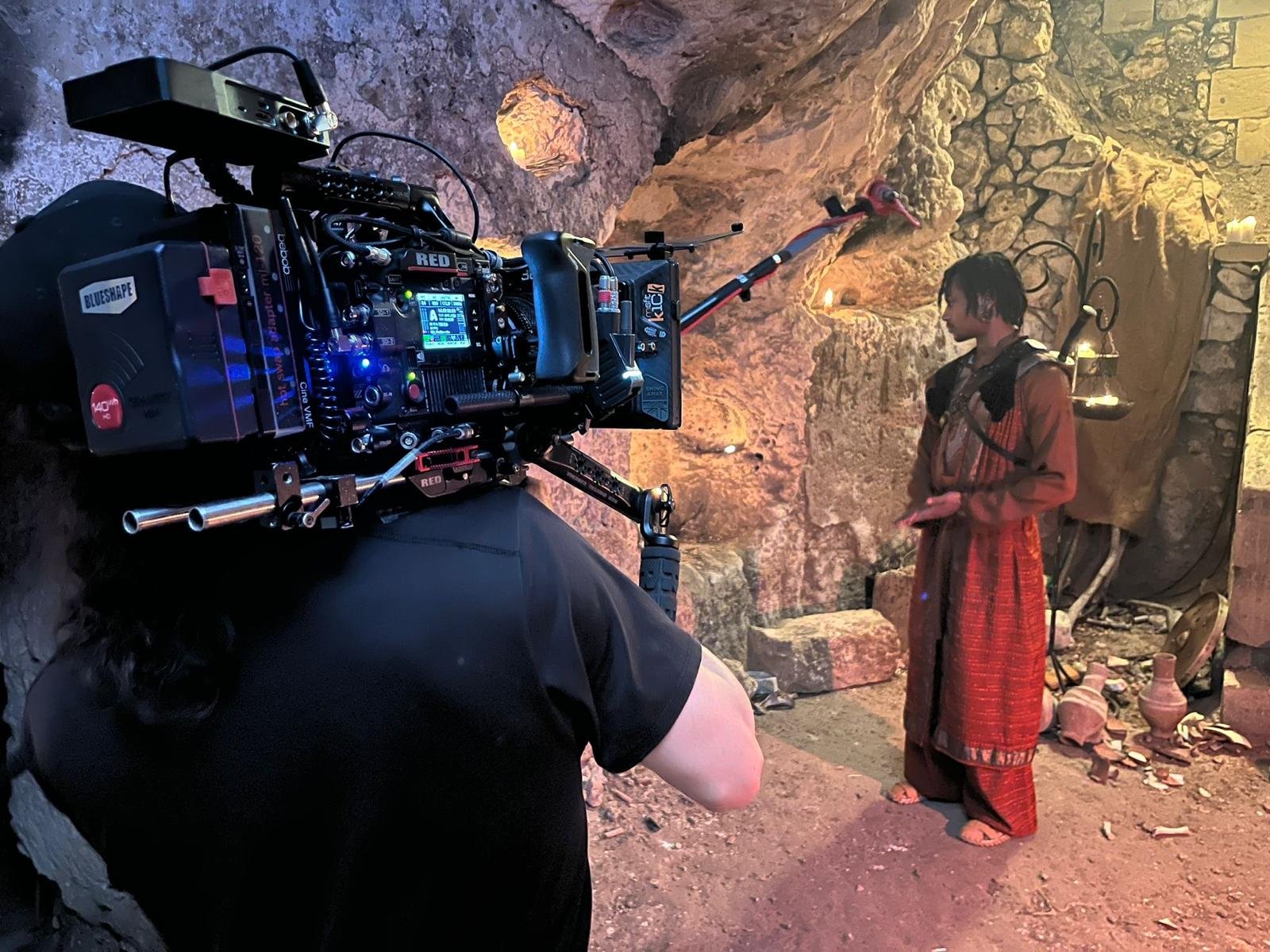 A behind-the-scenes shot from a film or TV production shows a camera on a tripod aimed at a woman dressed in ancient or traditional clothing, standing in a set designed to look like a cave or historical location with stone walls, pottery, and candle 