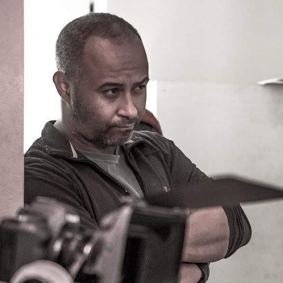 A man with short hair and a beard, wearing a dark jacket, looking down with his arms crossed in a room with white walls.