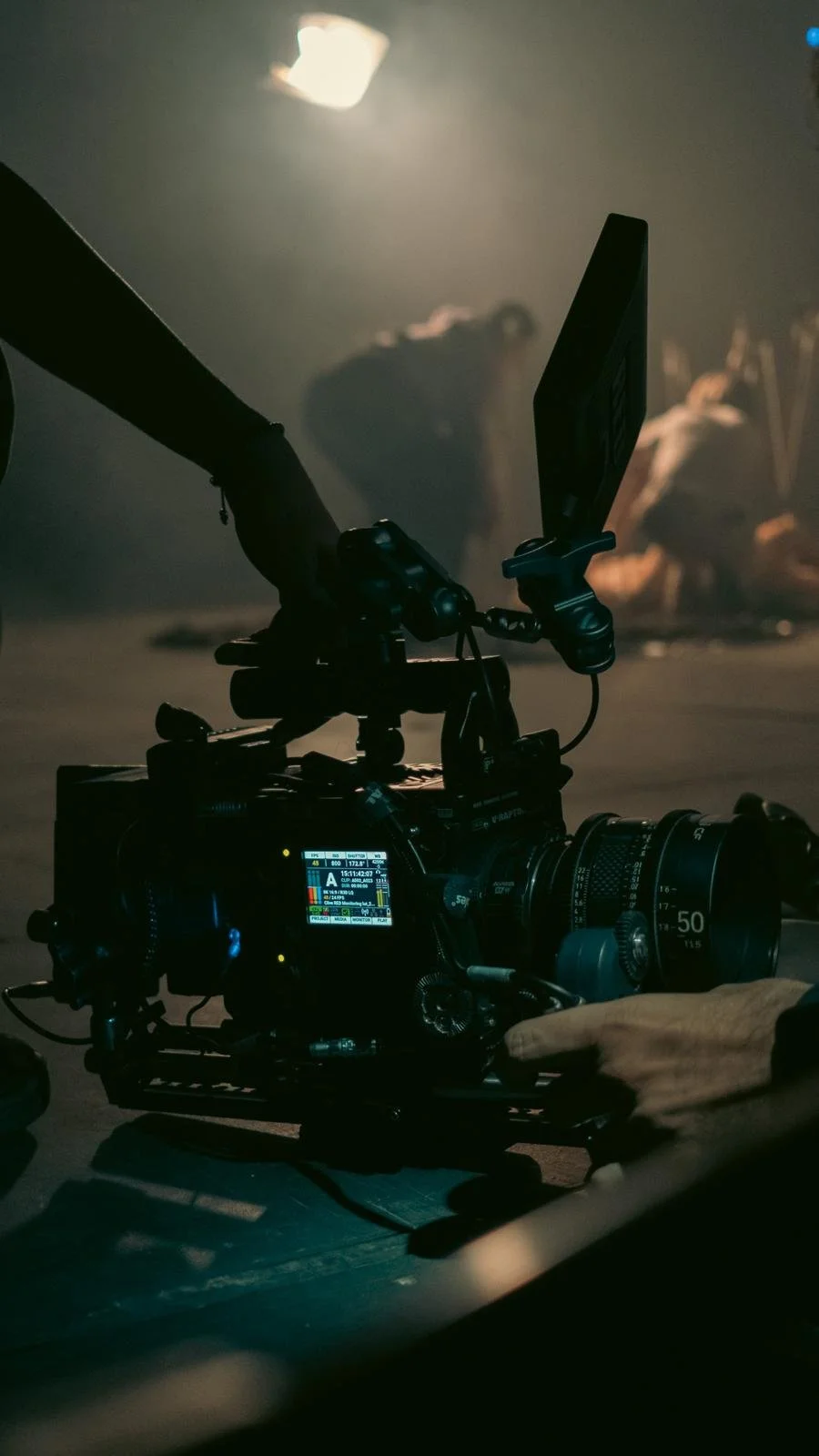 Close-up of a professional video camera with a monitor, situated in a dimly lit studio or set, with a blurred background showing a person and some equipment.