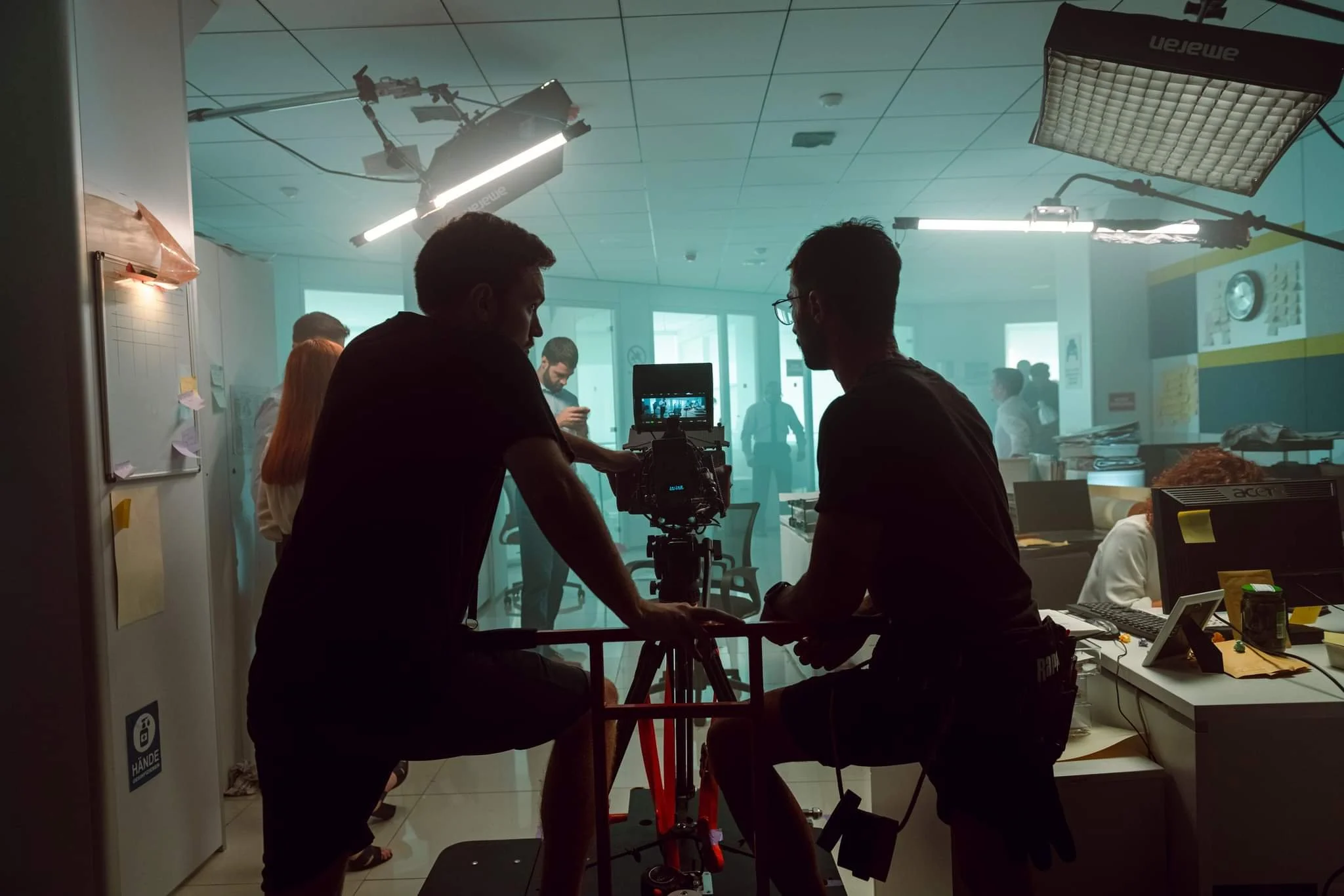 Two people working with a camera in an office, with a filming or lighting setup overhead and several colleagues in the background.