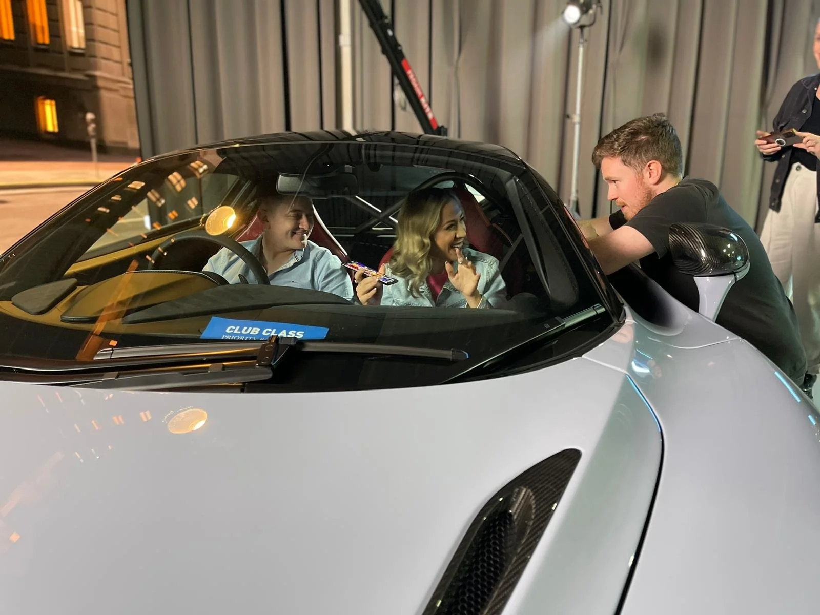 Two women in a sports car with a man leaning in and talking to them, during a nighttime event. The car has a 'Club Class' sticker on the dashboard.
