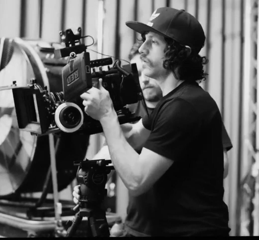 Black and white photo of a man with curly hair and a mustache operating a professional film camera, wearing a baseball cap, in a film set environment.