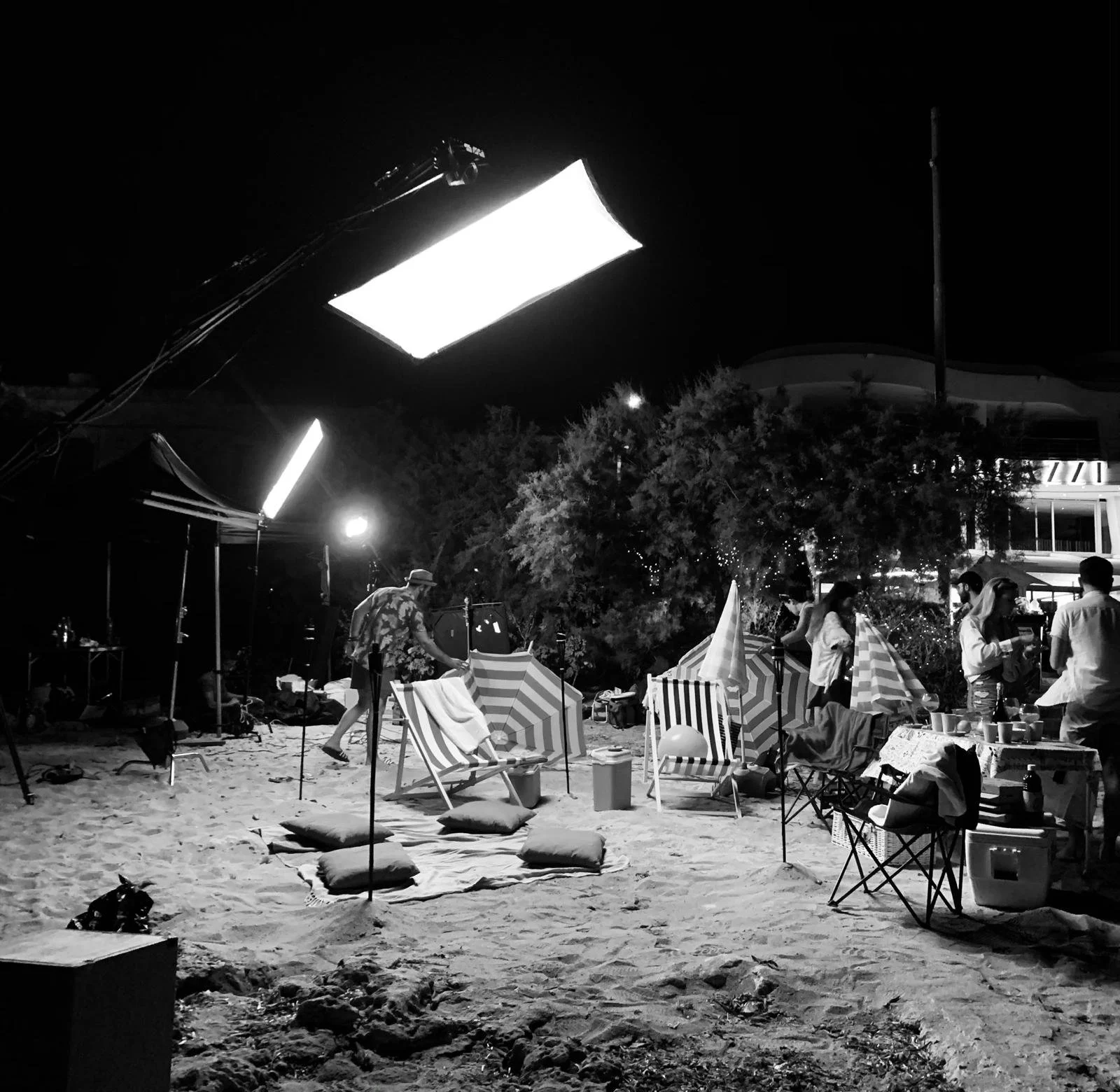 Night scene on a beach with people setting up or cleaning up, with umbrellas, chairs, and equipment, illuminated by large studio lights.