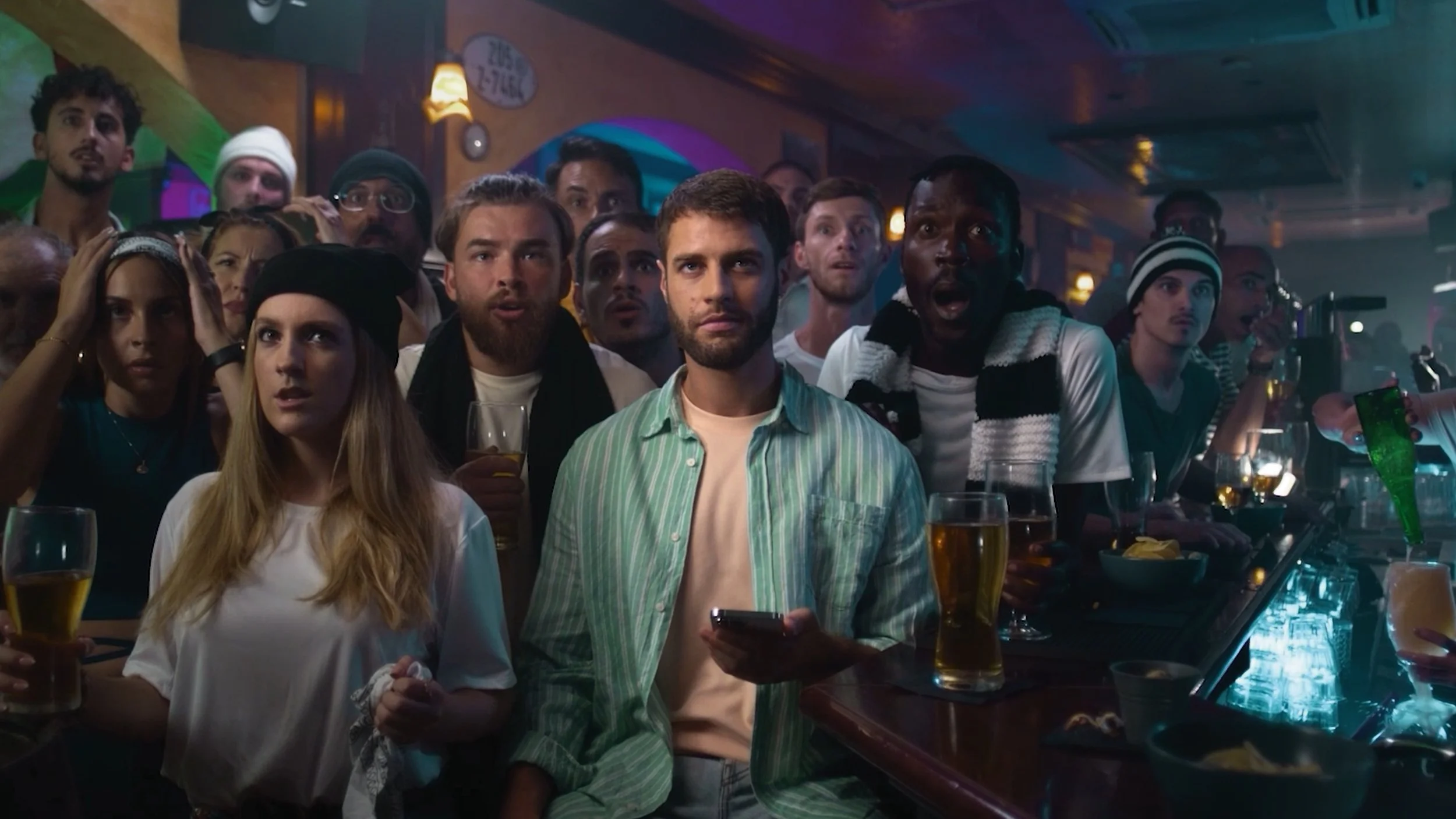 A group of diverse people gathered in a bar, watching something with varied expressions of surprise, excitement, and curiosity. They are surrounded by drinks and bar decor.