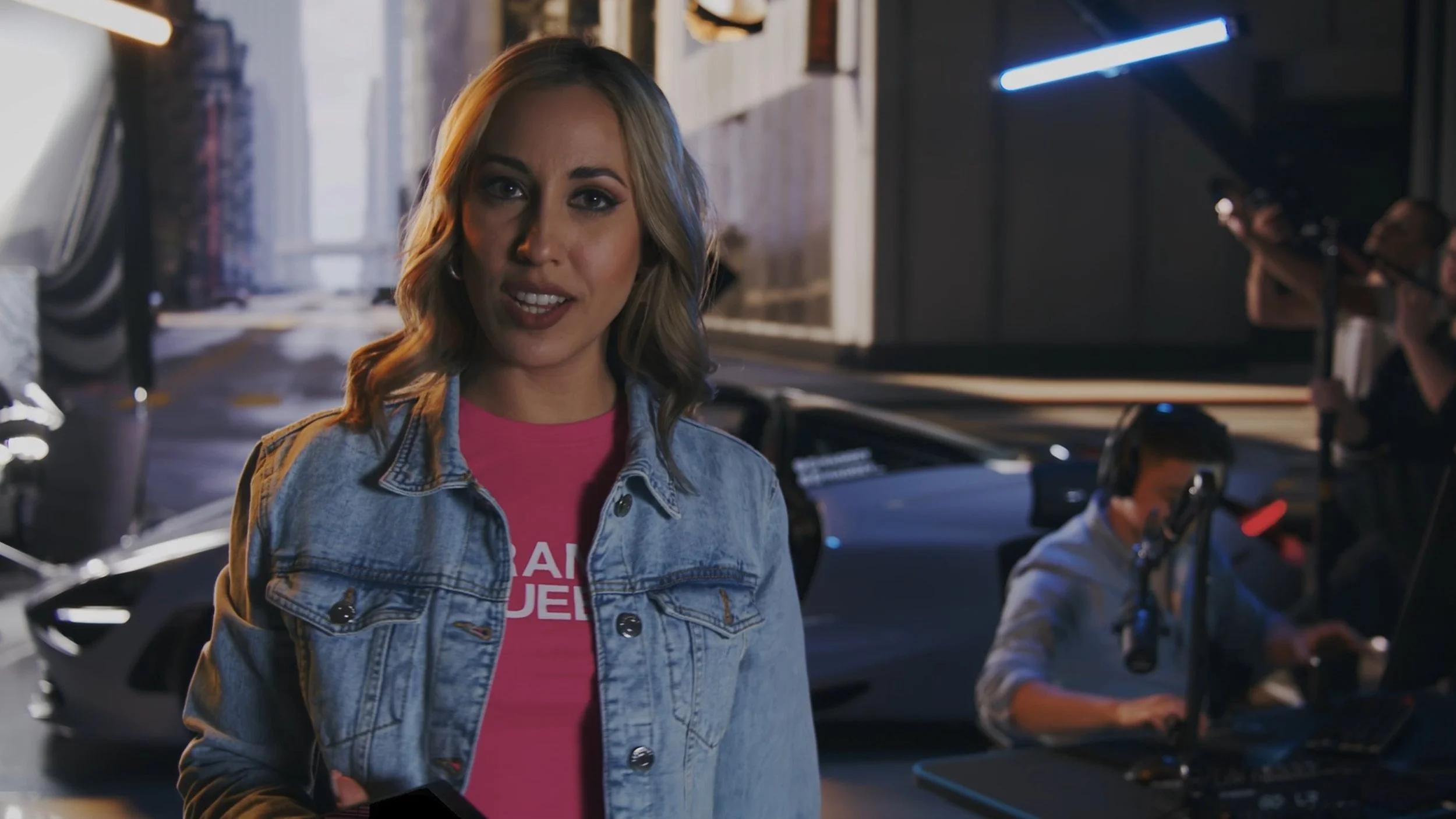 A woman with blonde hair, wearing a denim jacket and a pink shirt, standing in front of a filming set with cars and crew members working behind her.