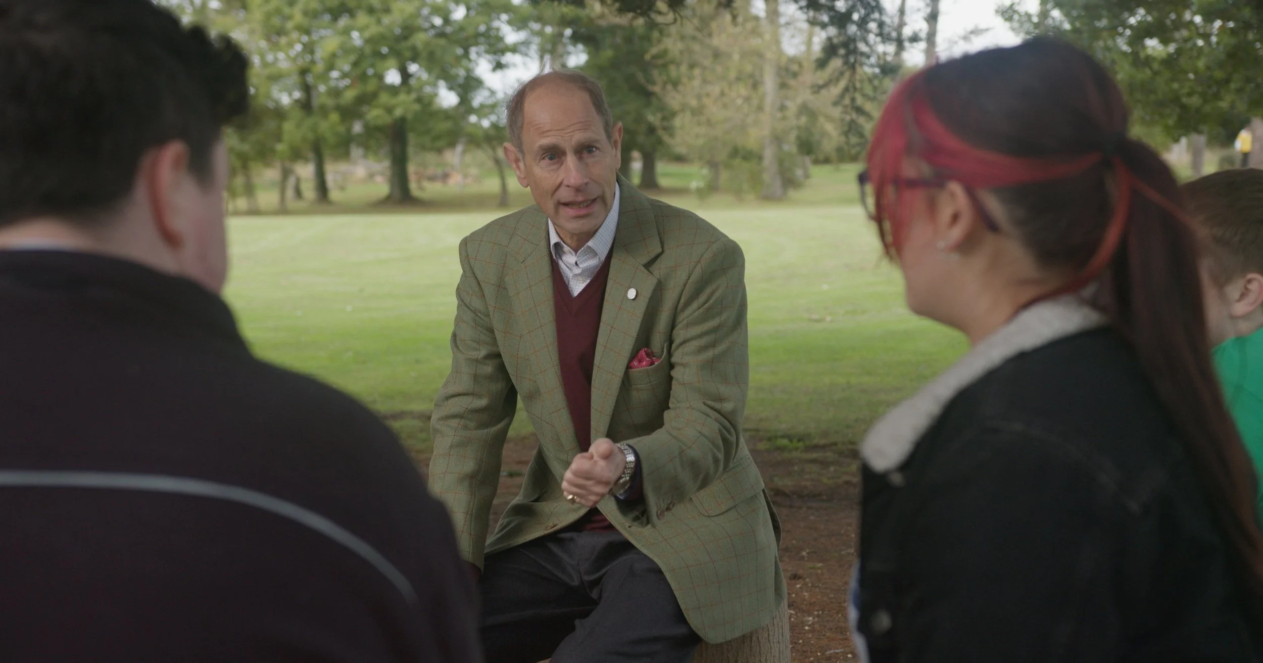 A man in a green checked blazer and red sweater shaking hands with a woman with red hair and glasses, while two other people look on in a park with trees in the background.