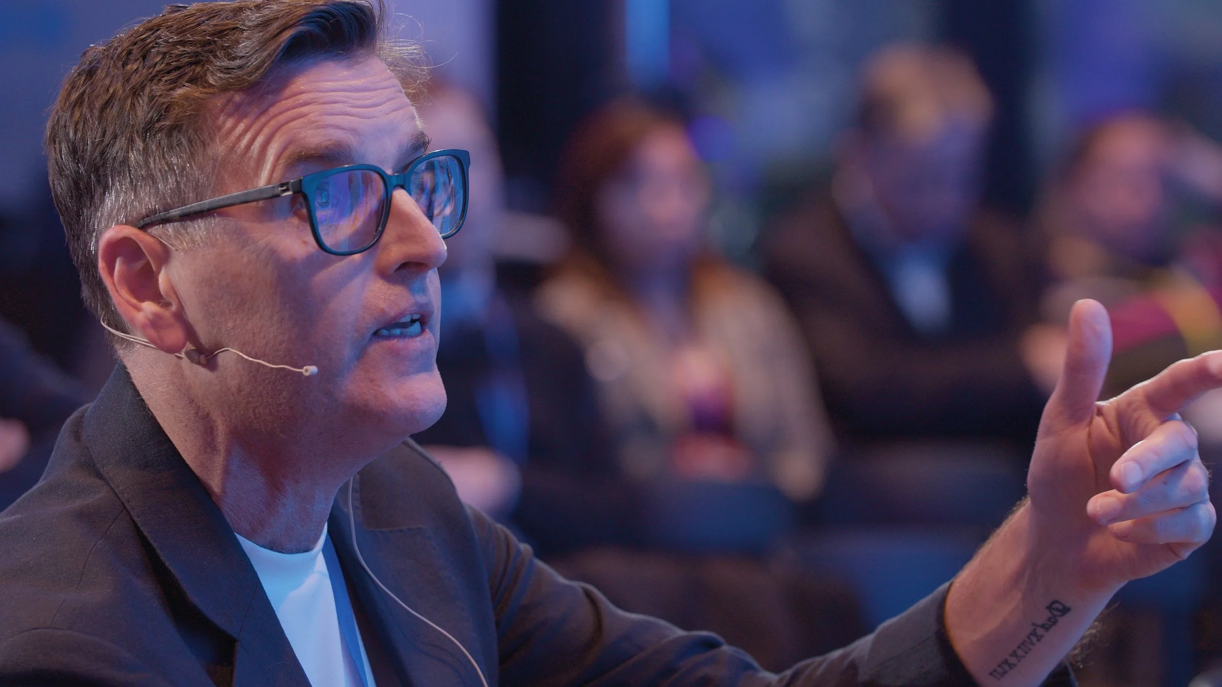 A man with glasses speaking at an event, wearing a jacket and shirt, in a dimly lit room with blurred audience in the background.