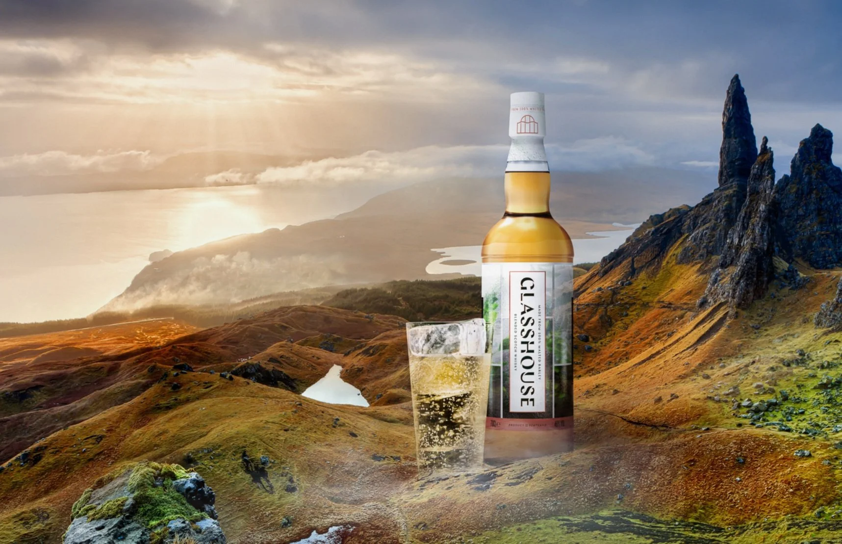 A bottle of Glashouse whiskey and a glass of whiskey with ice on a landscape of rolling hills, rocky formations, and a cloudy sky at sunset.