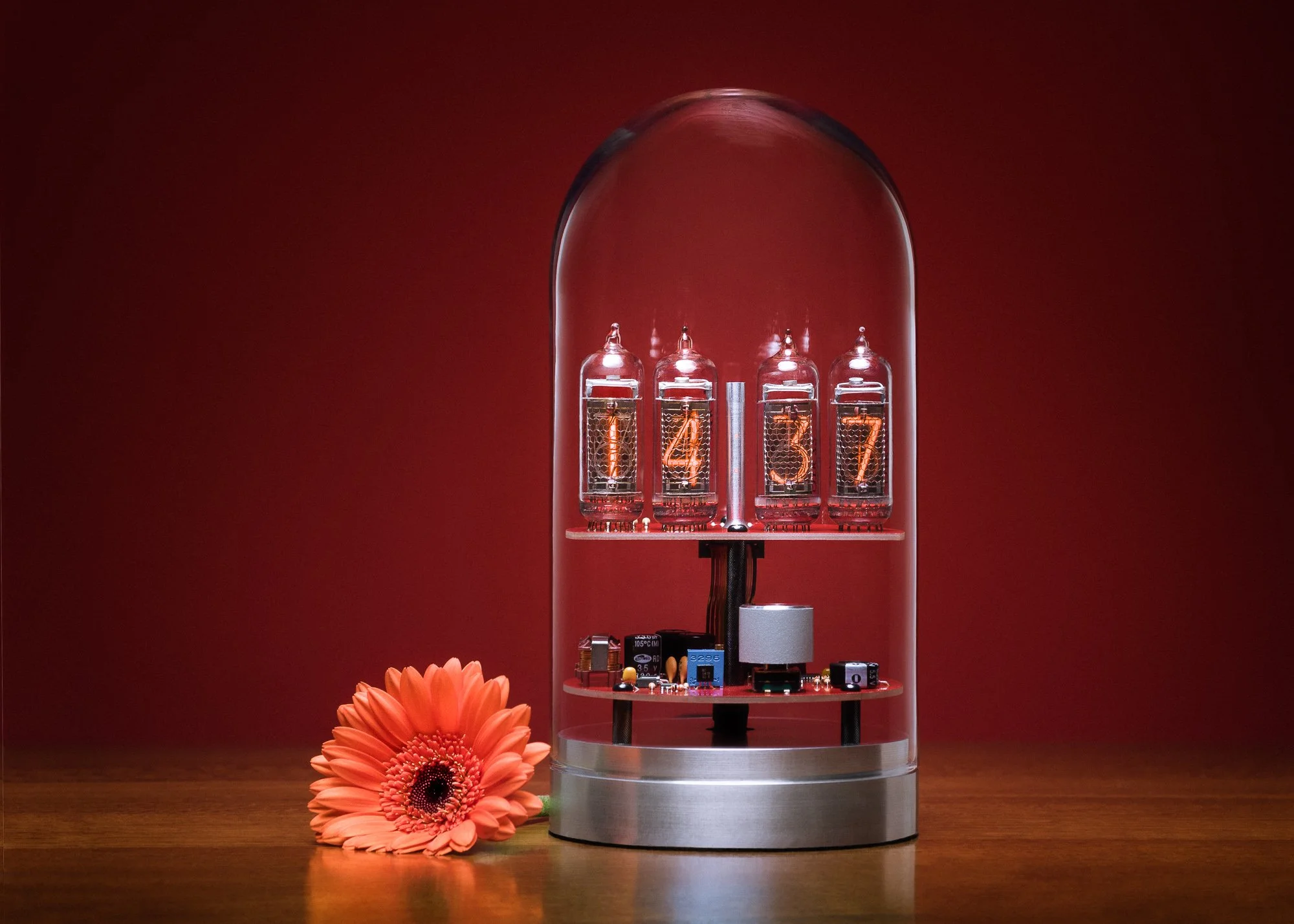 A modern clock with glowing N, 4, 3, 7 numerals inside a glass cover, placed on a wooden surface beside a bright orange gerbera flower, against a red background.