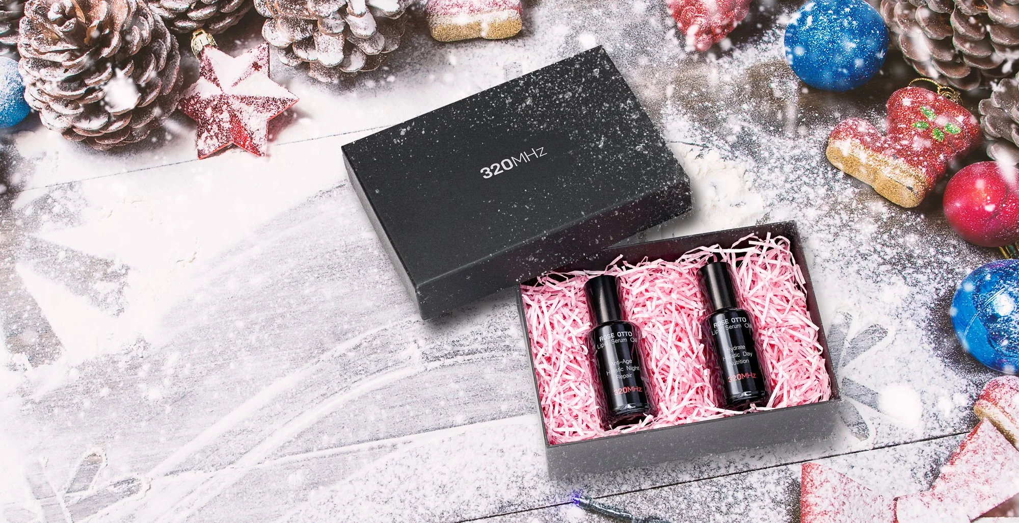 A Christmas-themed scene with pine cones, decorated ornaments, some sprinkled with artificial snow, and two small bottles of lip serum in a gift box filled with pink shredded paper. The background features a white wooden surface dusted with artificia