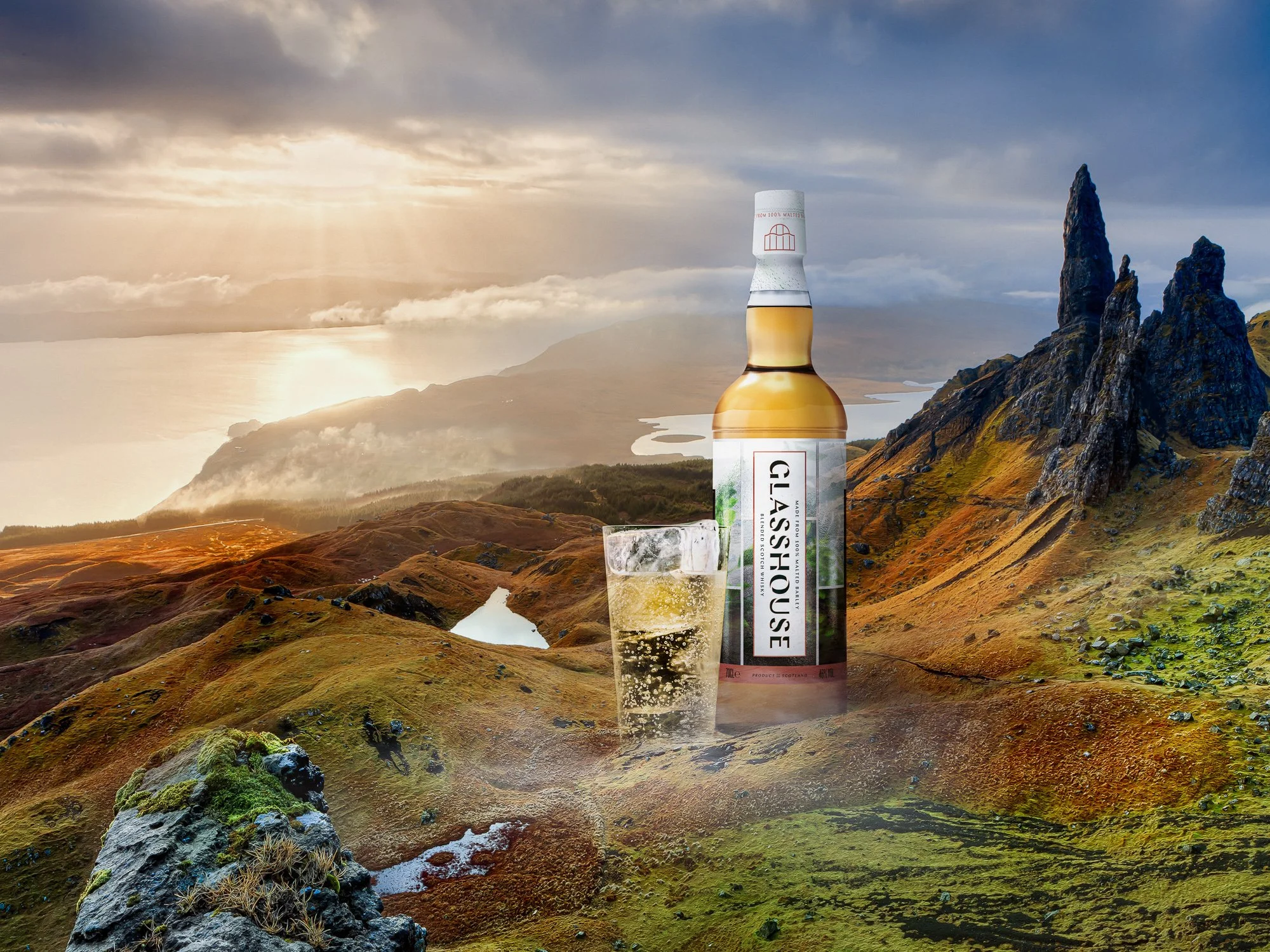 A scenic landscape of mountains and lakes at sunset or sunrise with a giant bottle of Glenfiddich Scotch whisky and a glass filled with ice and whisky placed in the foreground.