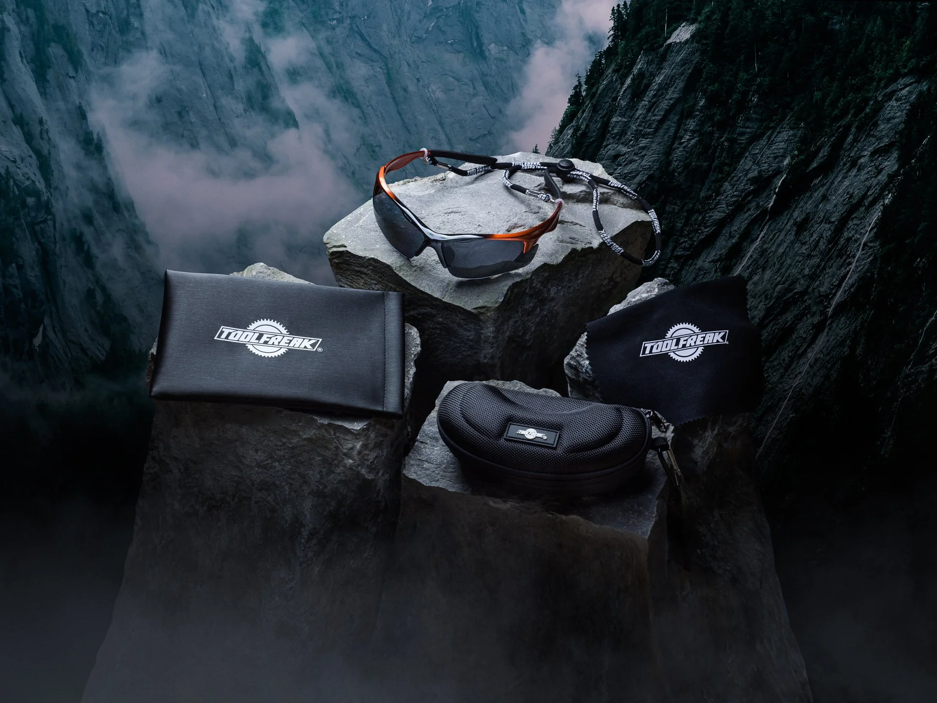 Climbing gear including sunglasses, a headlamp, and bags resting on rocks at a mountain edge with a deep canyon and fog in the background.