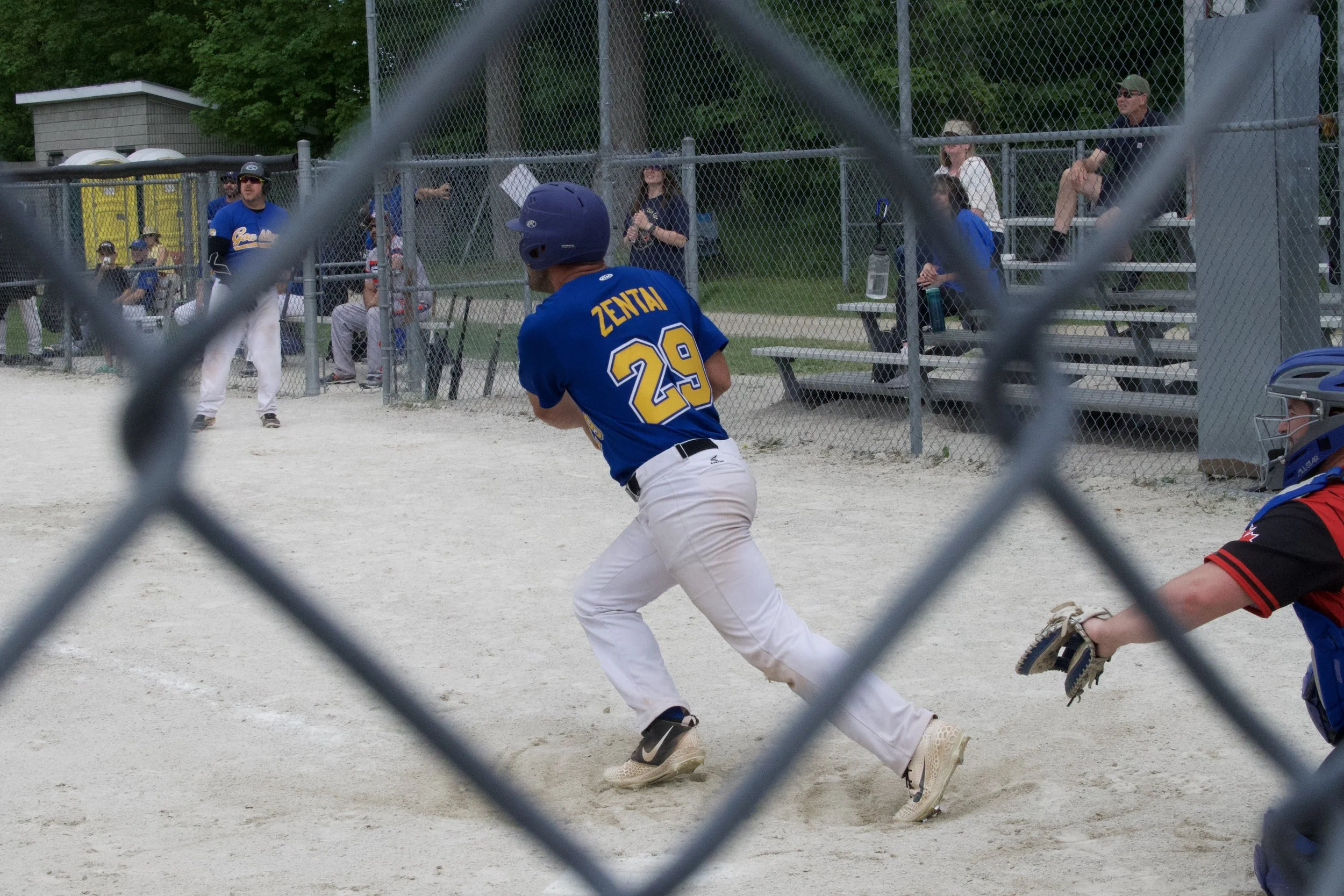 A baseball player wearing a blue jersey with the name ‘ZENTAI’ and number 29 is running on the field, viewed through a chain-link fence. In the background, people are sitting on bleachers watching the game at the Vasey, Ontario baseball diamond.