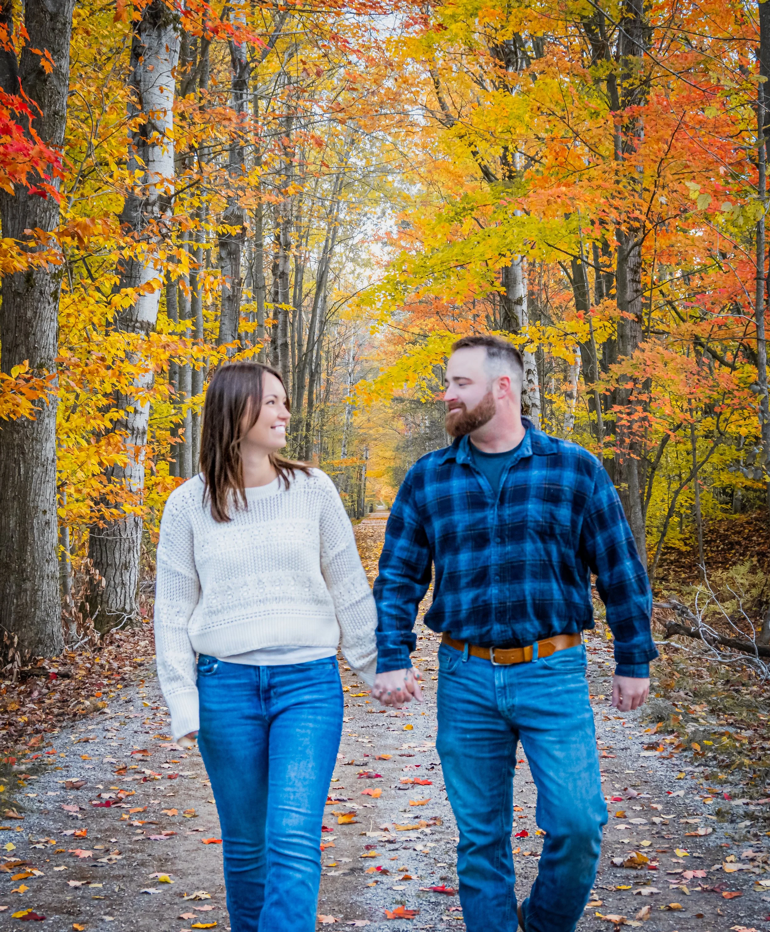 A young couple holding hands and walking together in a park during autumn, surrounded by trees with colorful fall leaves in shades of yellow, orange, and red.