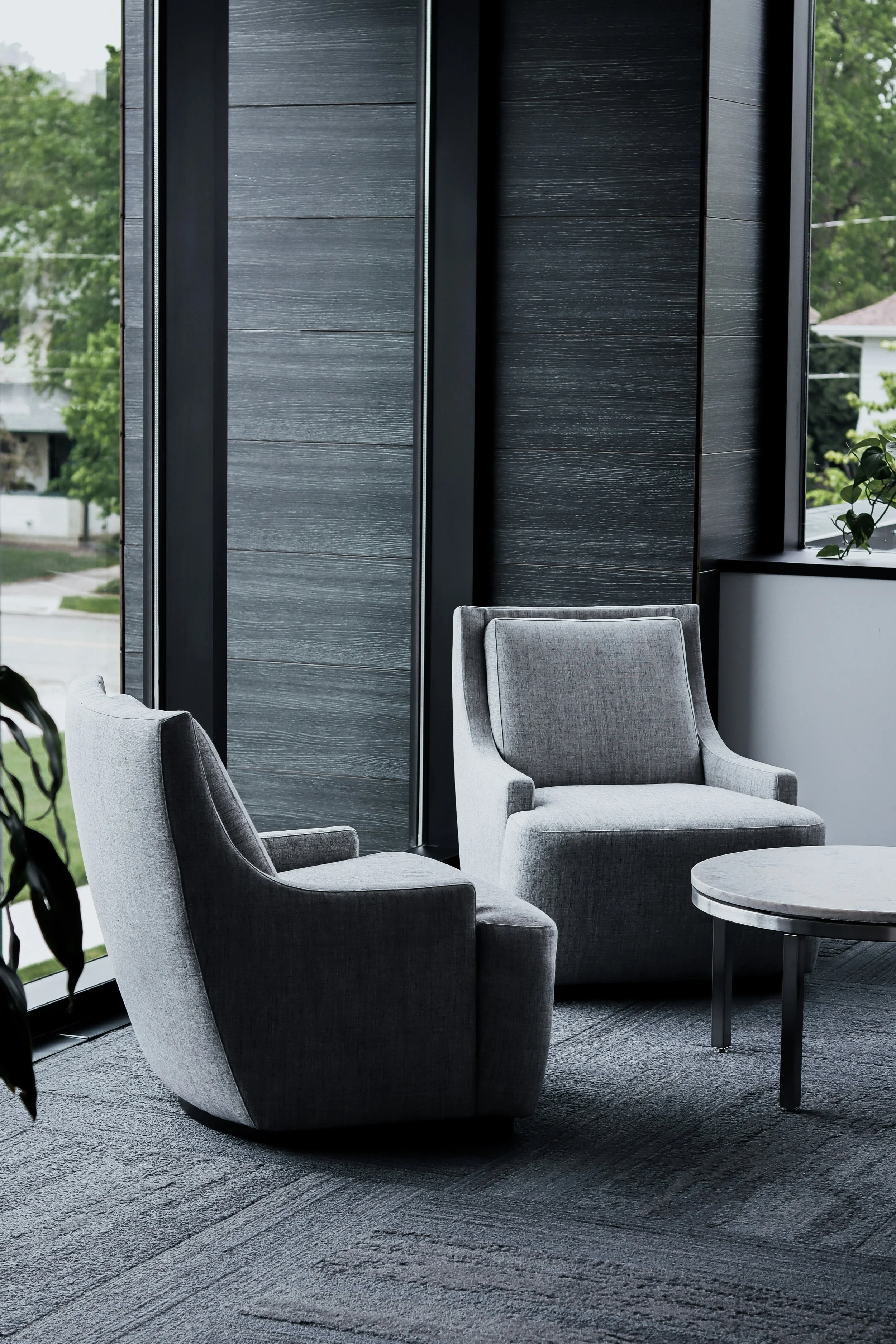 Modern interior lounge with gray armchairs, a small round table, dark wooden wall panels, and large windows with a view of green trees outside.