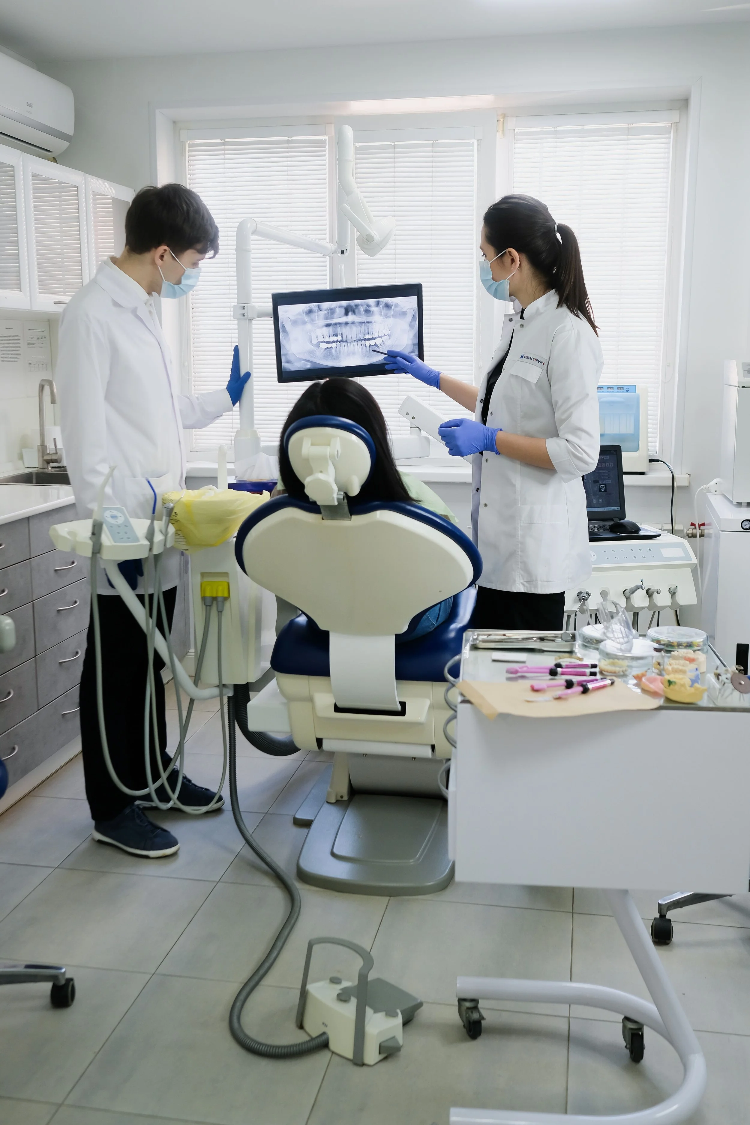 Dentists and a patient in a dental clinic examining a panoramic dental X-ray. The dentists are wearing face masks and gloves, and the patient is seated in a dental chair.