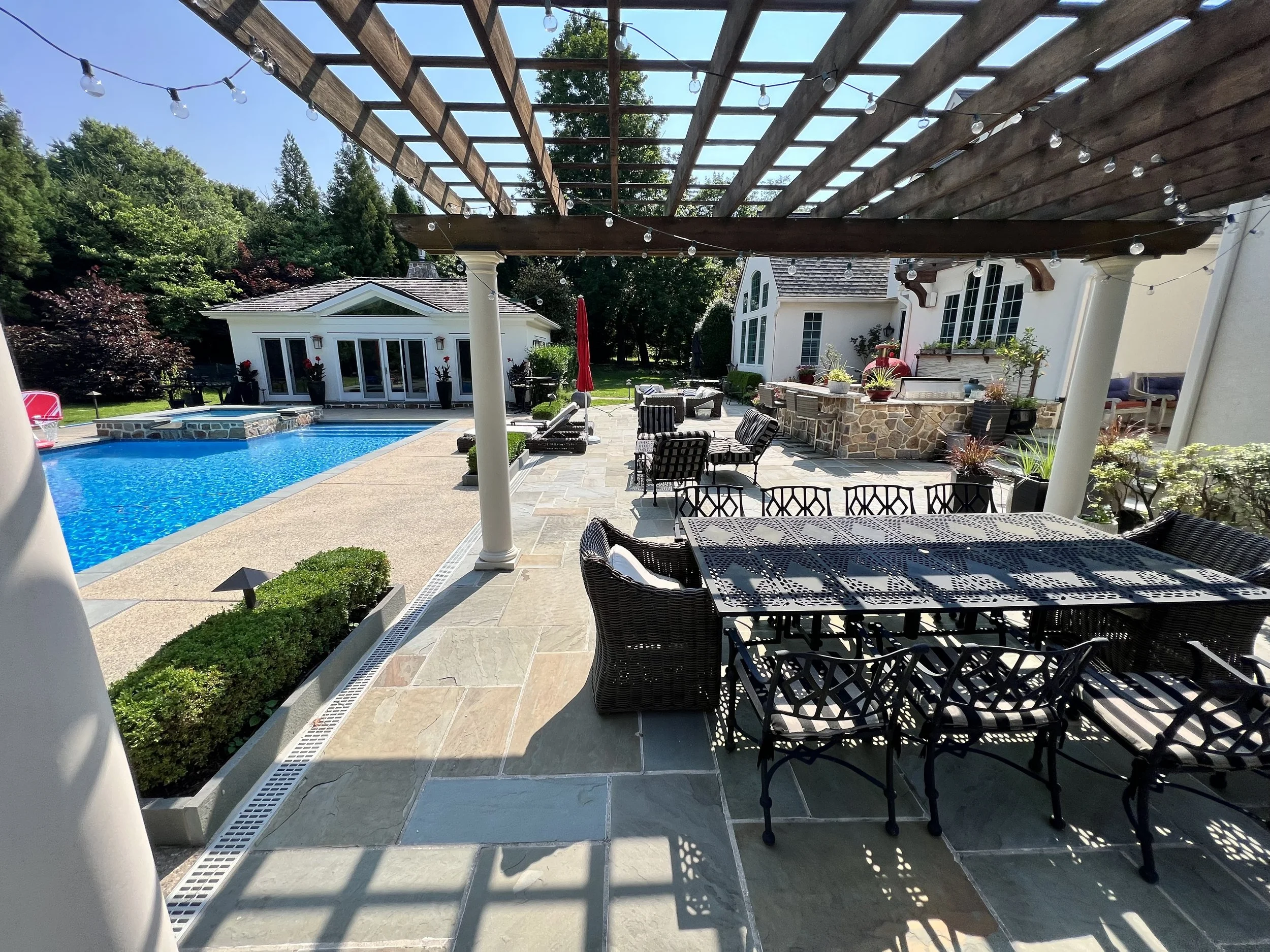 View of a backyard patio area with outdoor furniture, a swimming pool, a small building with glass doors, and a spacious deck covered by a wooden pergola with string lights. There are lush green trees and a clear sky in the background.