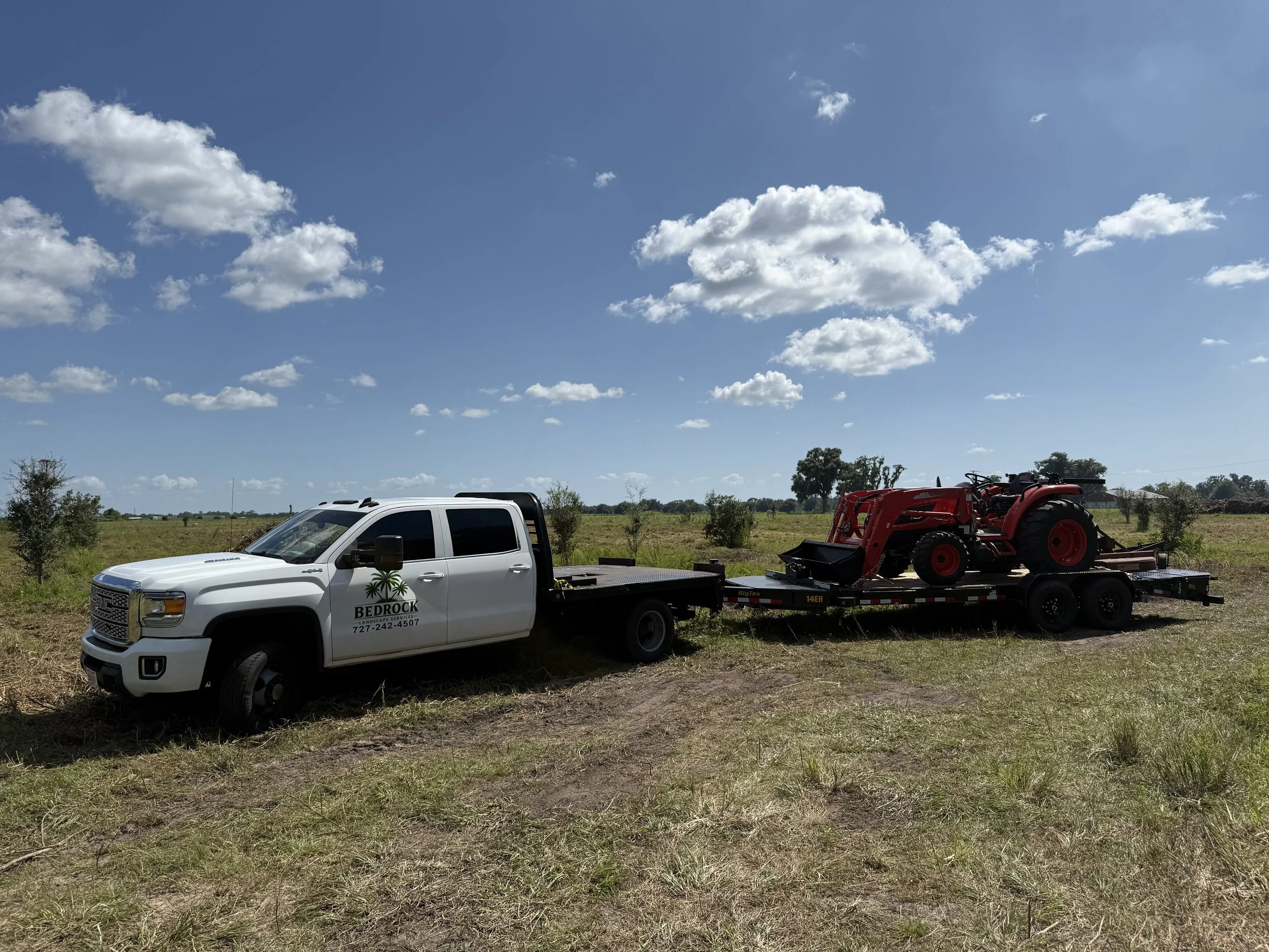 A white pickup truck with a Bedrock Landscape Services logo on the door, carrying a red tractor on its flatbed in a rural area with scattered trees and a blue sky with clouds.