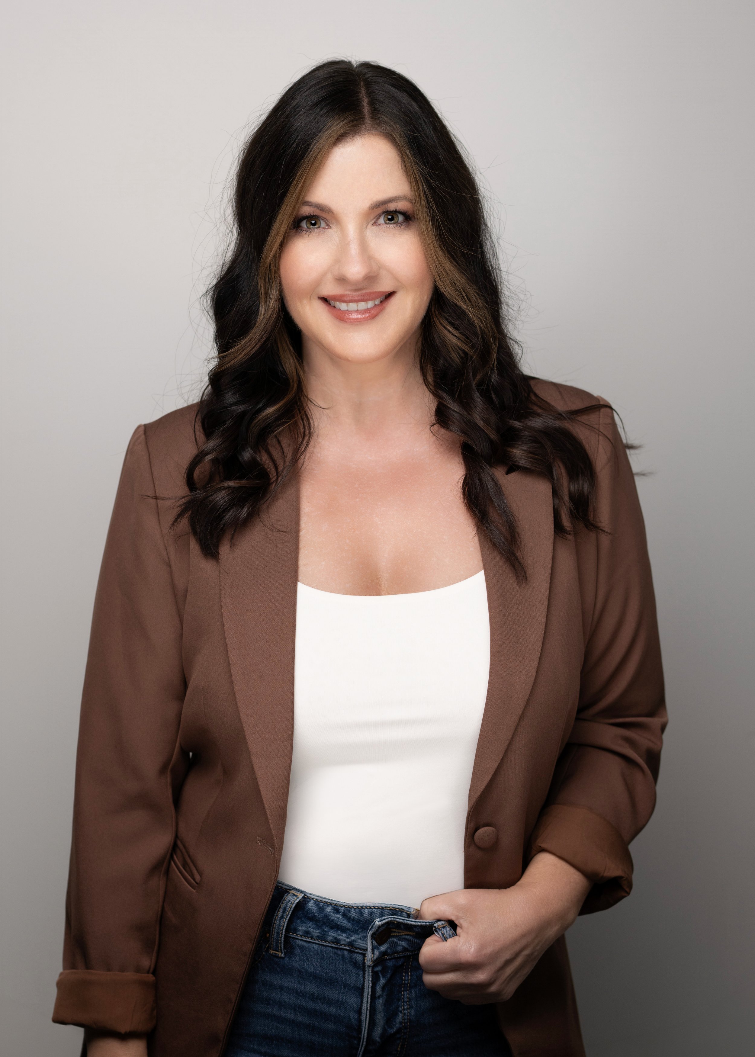 A woman with long dark hair, wearing a brown blazer over a white top, smiling and standing against a plain light gray background.