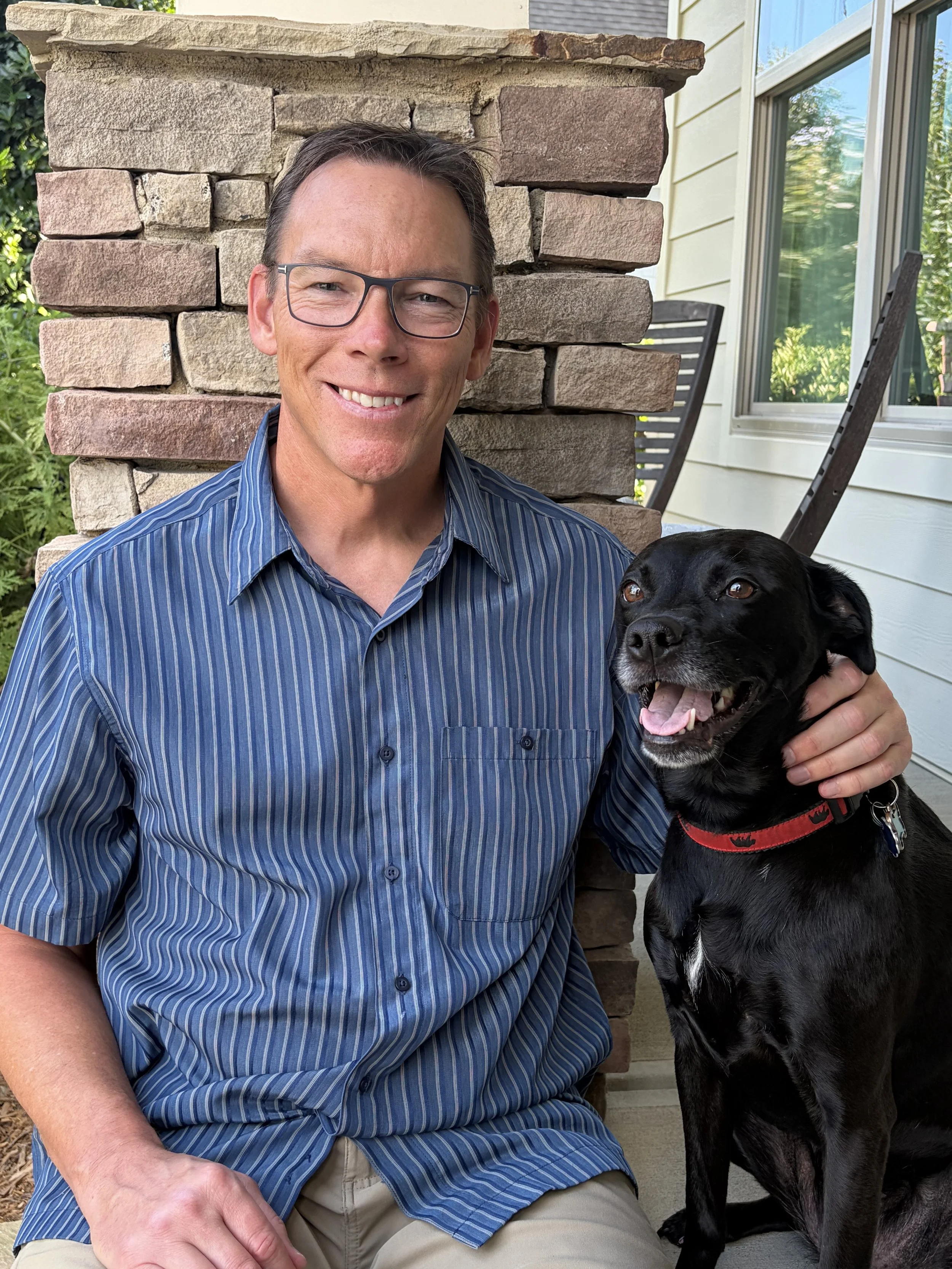 A man with glasses sitting outside on a porch next to a black dog with a red collar, both smiling and looking at the camera, with a stone wall and window in the background.