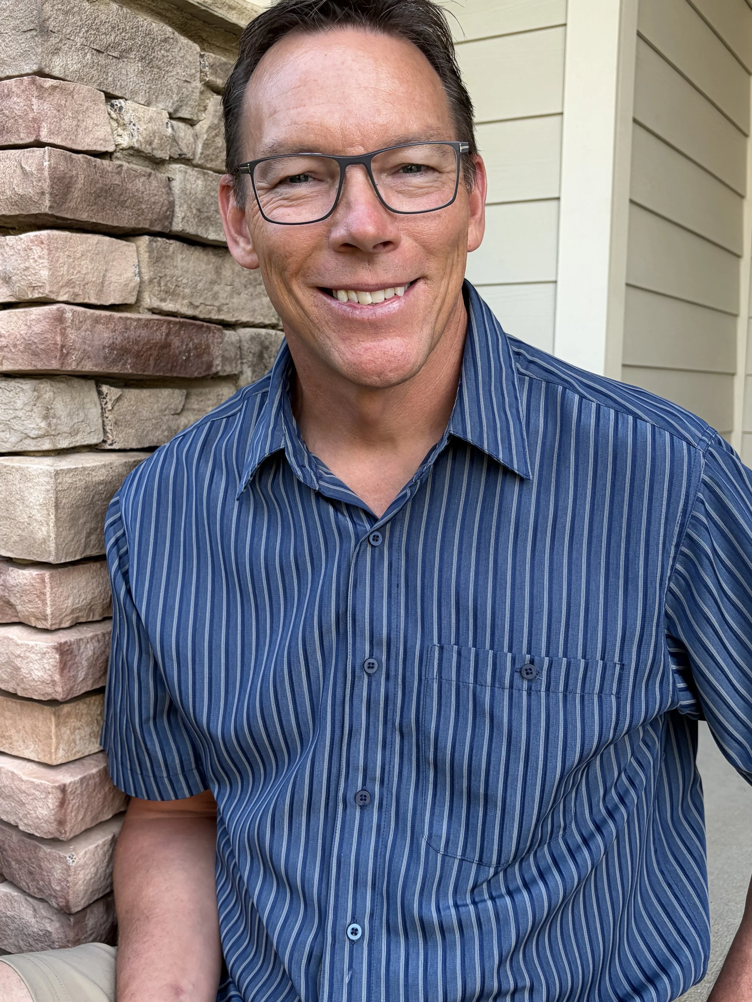A smiling middle-aged man with glasses wearing a blue striped shirt, sitting outdoors near a stone wall and a beige house siding.