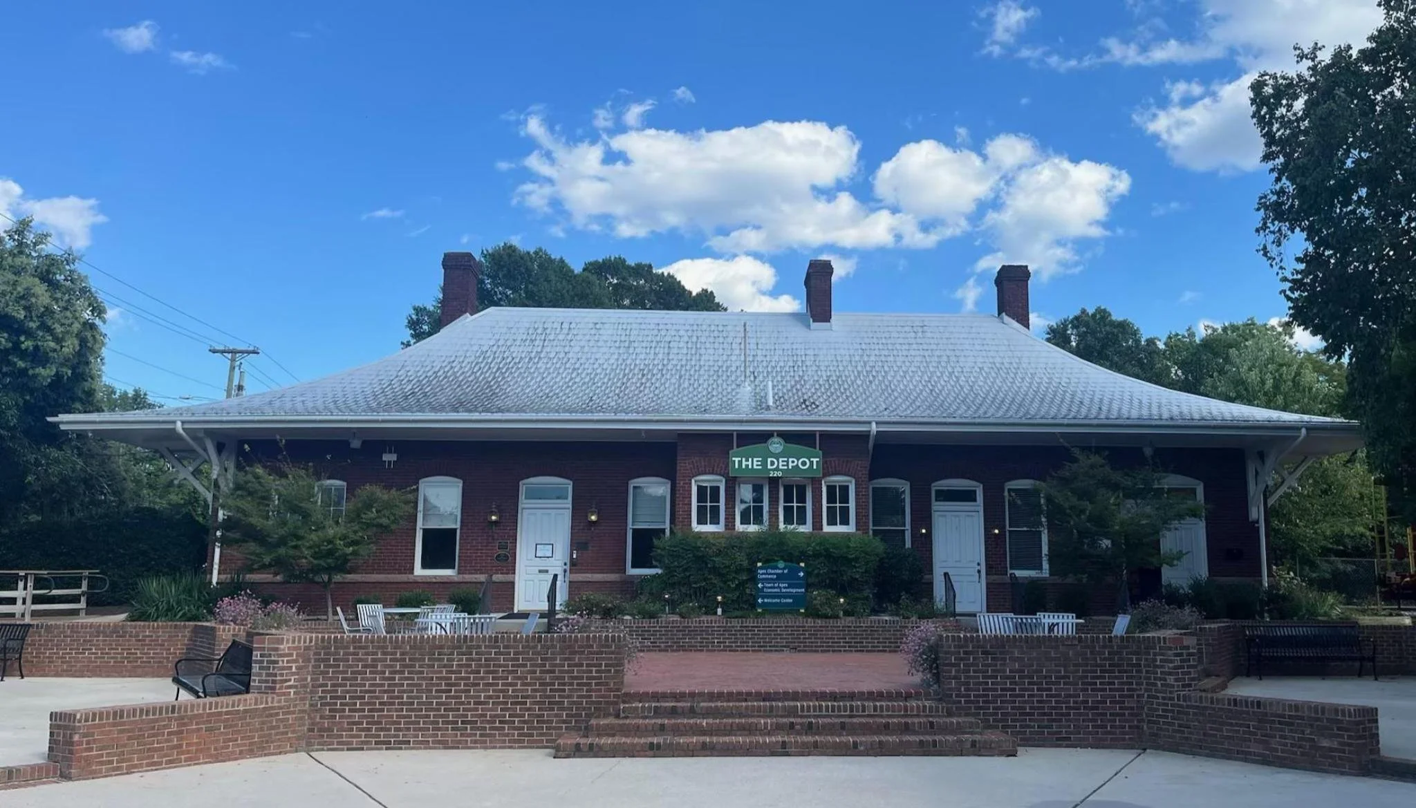 A brick building with a sign reading 'The Depot' under a blue sky with clouds, surrounded by trees and outdoor seating.