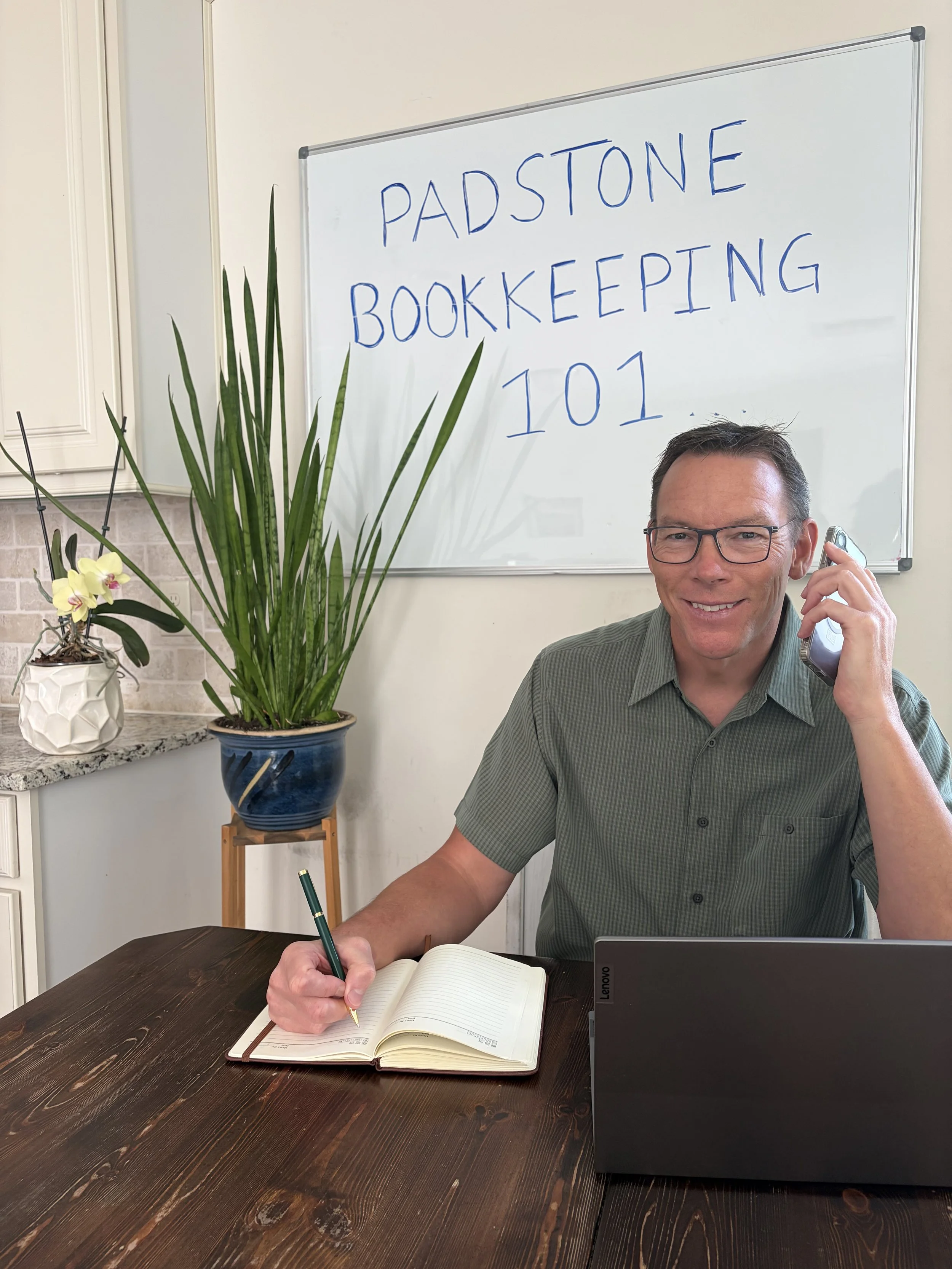 A man with glasses sitting at a dark wood table, talking on a cordless phone, writing in a notebook, with a partially open laptop in front of him. Behind him is a whiteboard with 'PADSTONE BOOKKEEPING 101' written on it. To his left, there is a tall potted plant and a white orchid in a decorative white vase.