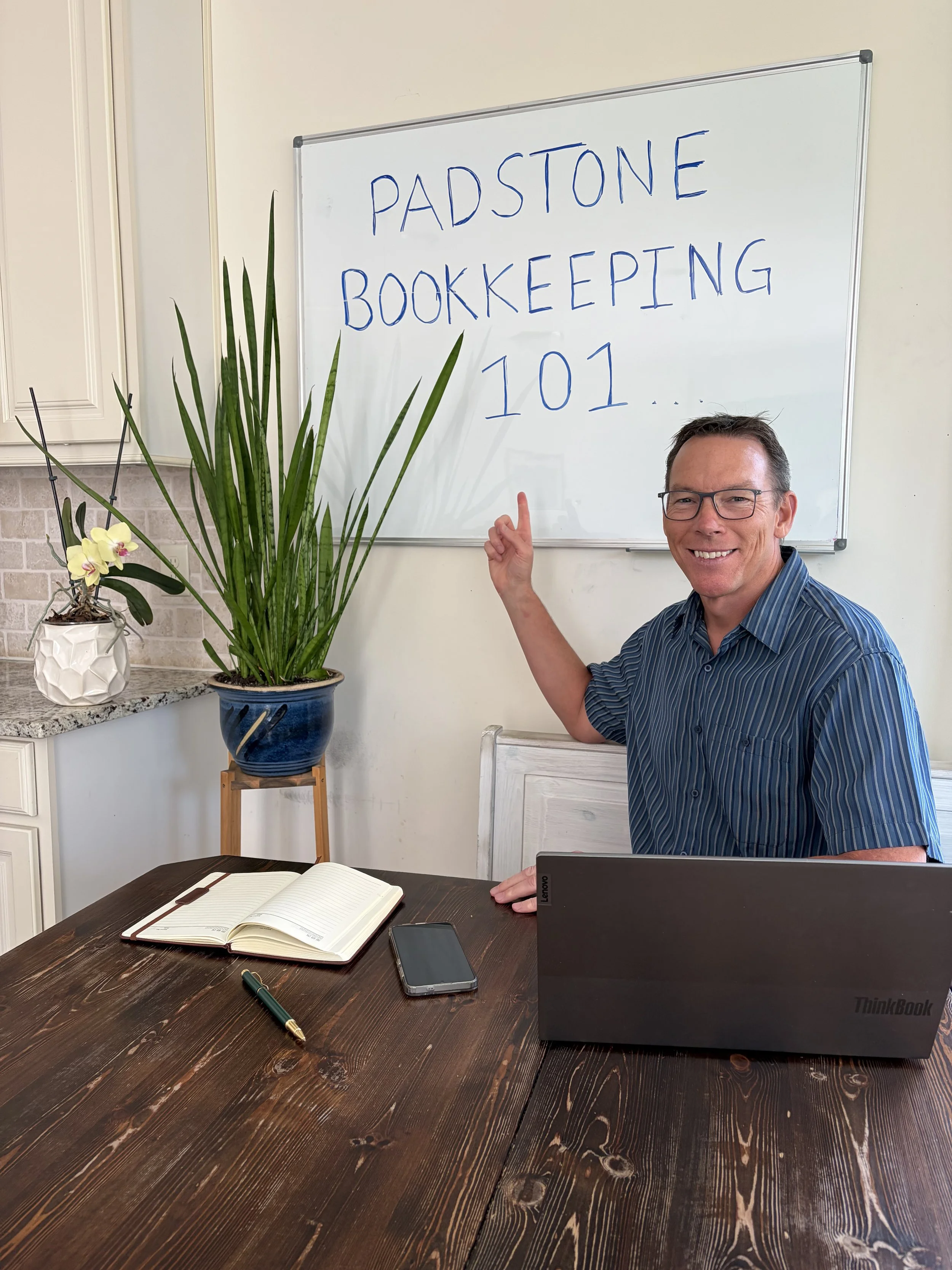 A smiling man wearing glasses and a blue striped shirt points to a whiteboard that reads 'Padstone Bookkeeping 101.' He is seated at a dark wooden table with an open notebook, a pen, a smartphone, and a laptop. There is a tall green plant and a potted orchid on the counter against a white wall.
