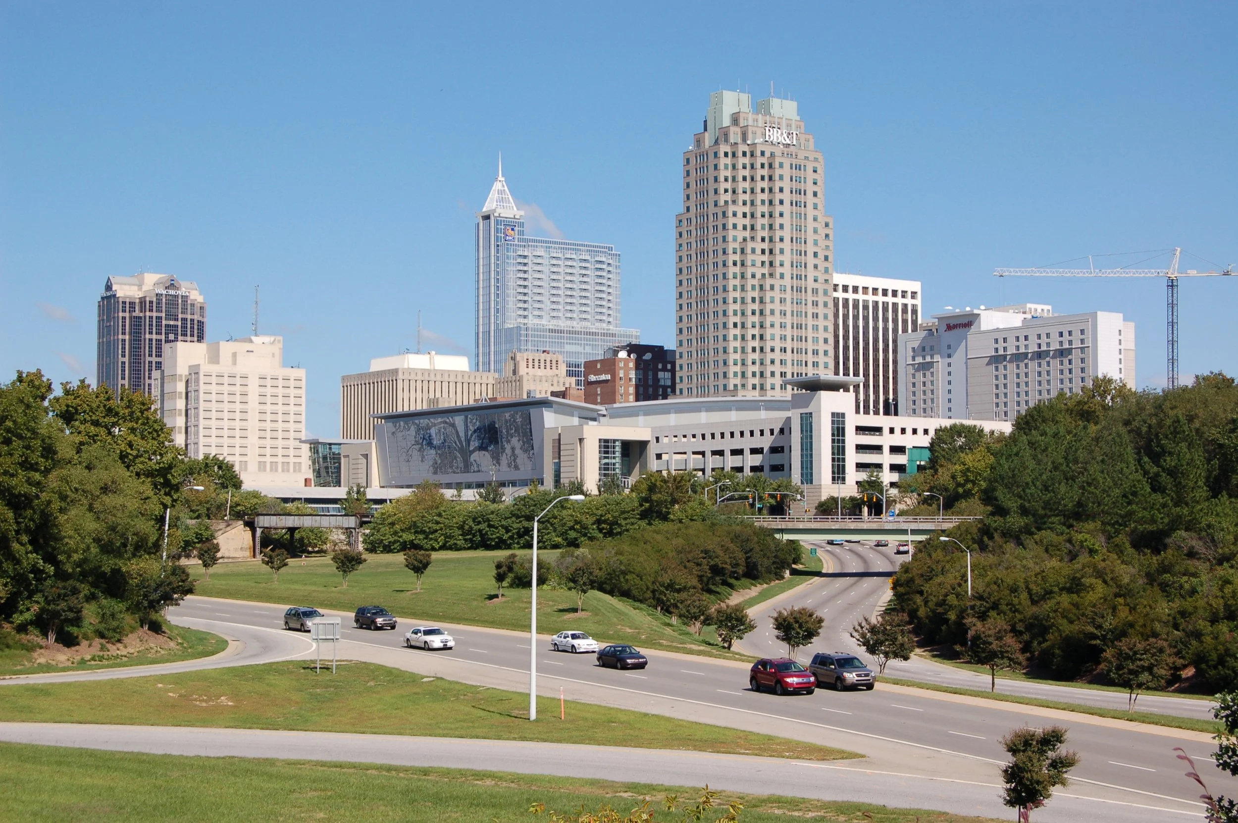 Downtown skyline with tall buildings, green trees, and a multi-lane road with cars under a clear blue sky.