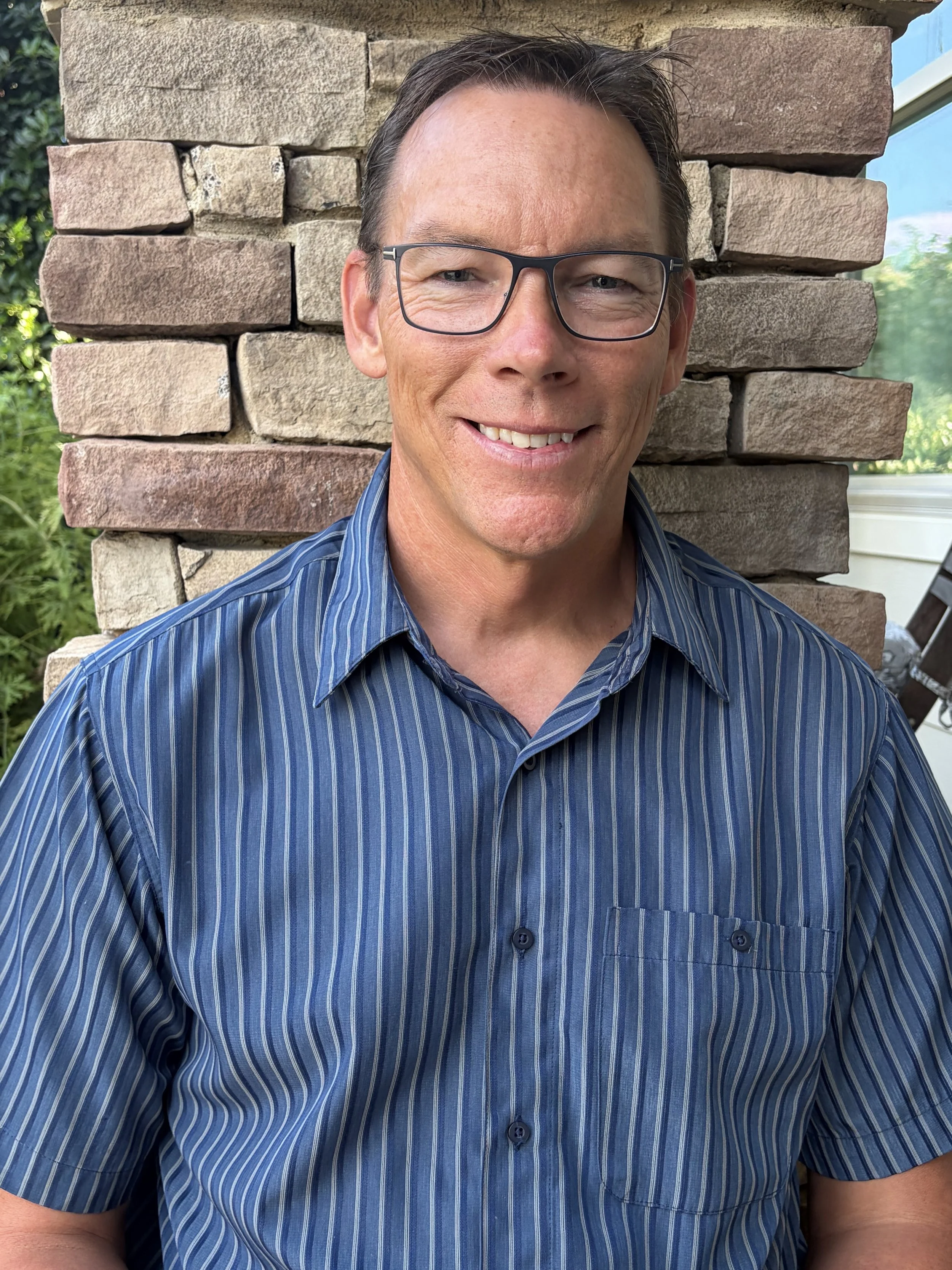 A man with glasses wearing a blue striped shirt, smiling, standing in front of a brick wall with some greenery and a window in the background.