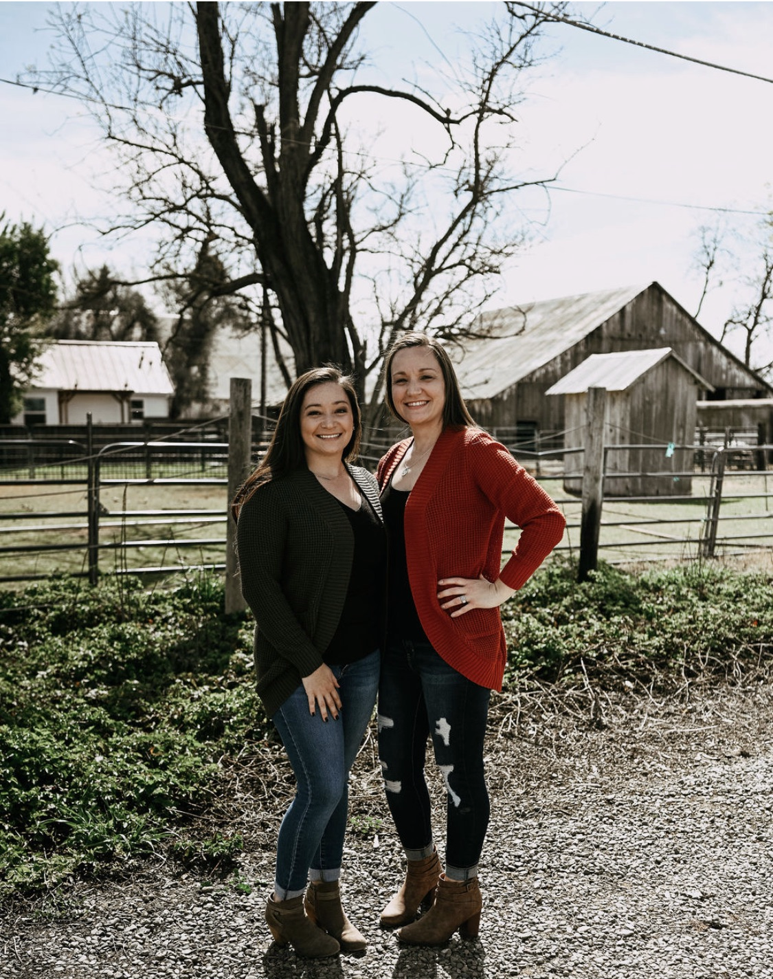 Two women standing outdoors, smiling, in front of a wooden fence with farm buildings in the background, a large leafless tree, and a partly cloudy sky.