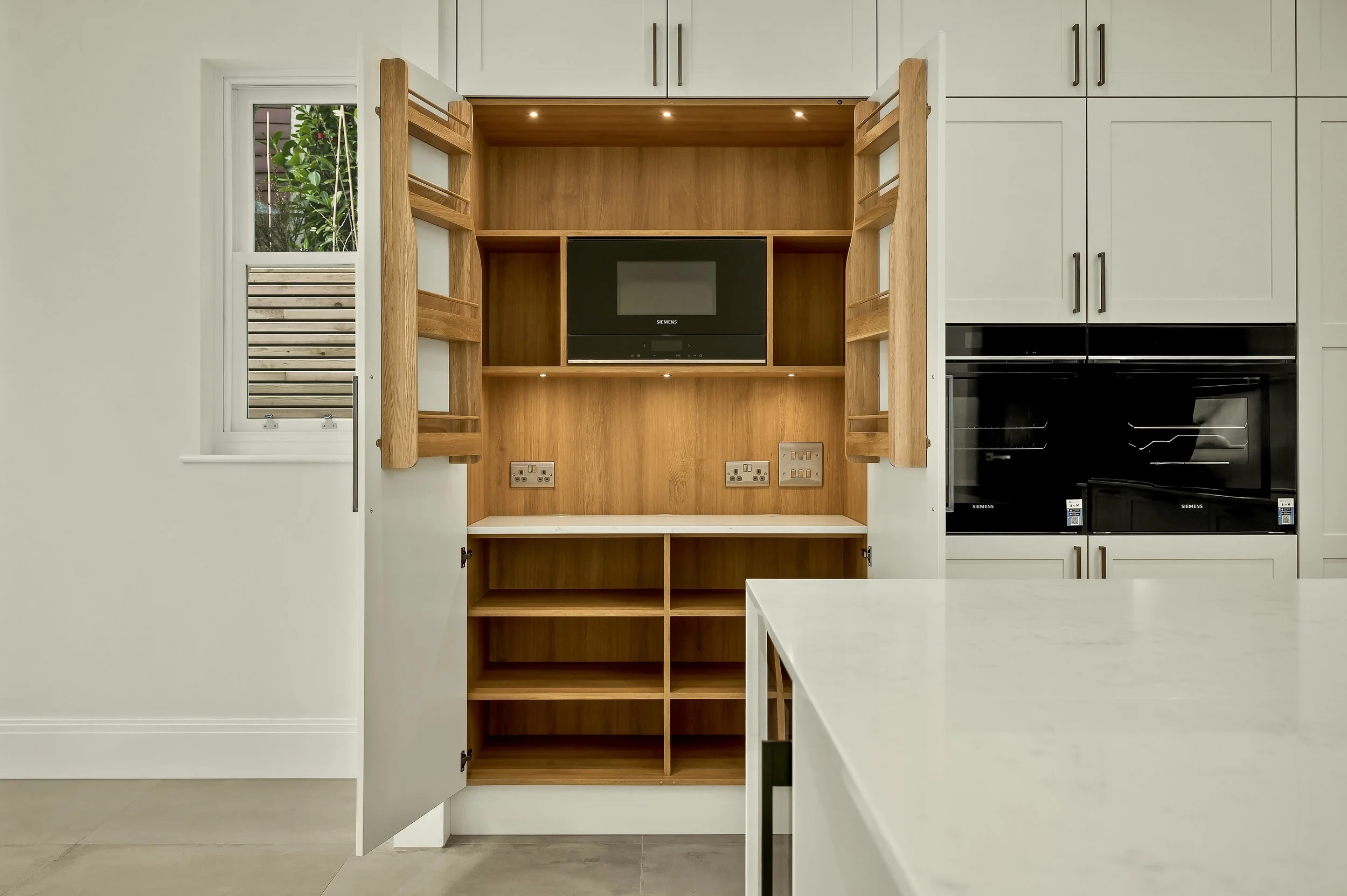 A kitchen with a white countertop in the foreground, a built-in wooden cabinet with open shelves and a black Siemens microwave in the center, white cabinetry above, and a window with a view of greenery on the left side.