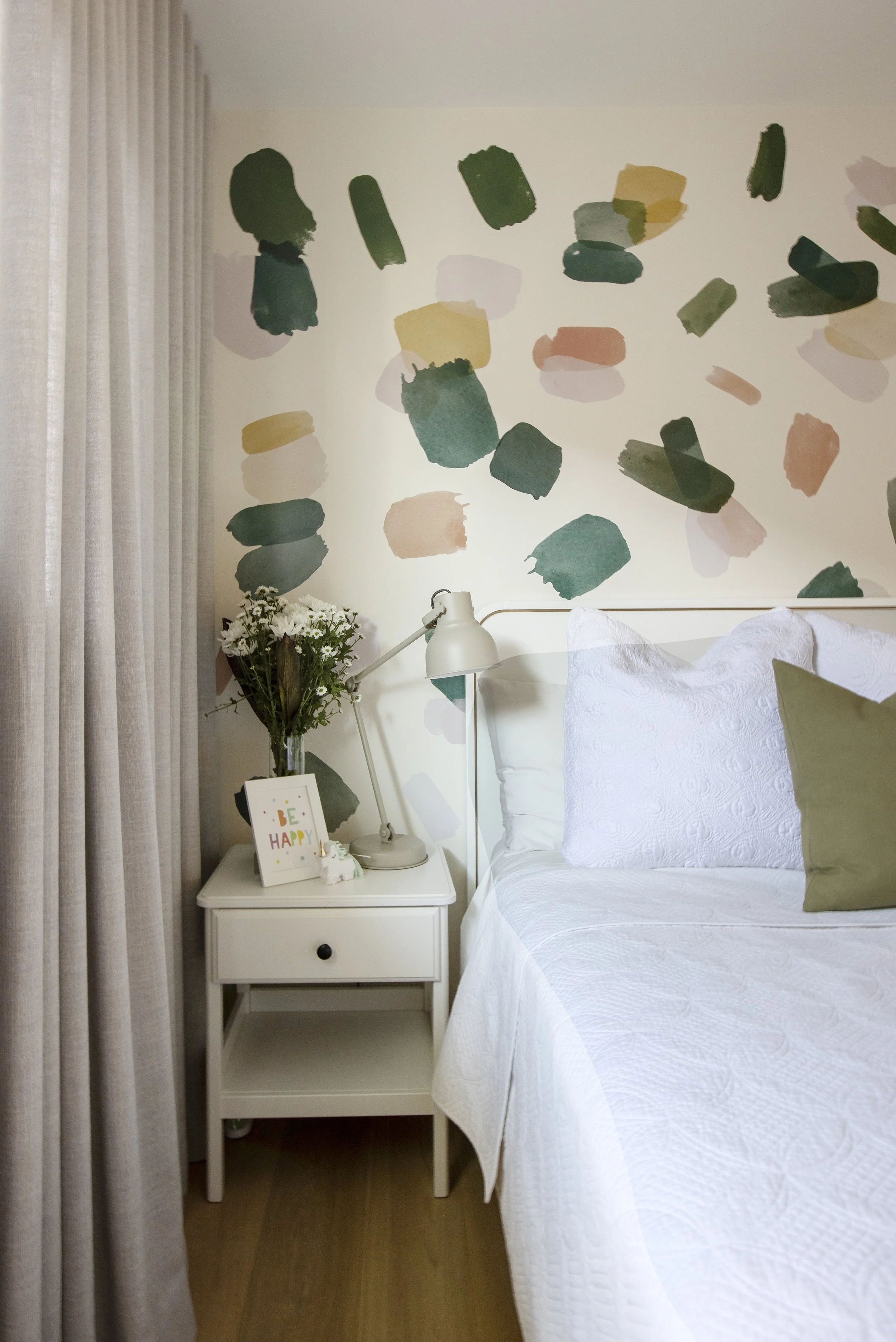 A bedroom with a white bedspread, a green pillow, a white bedside table with a black knob drawer, a white lamp, a bouquet of white daisies, a framed 'Be Happy' quote, and a colorful abstract wall art above the bed.