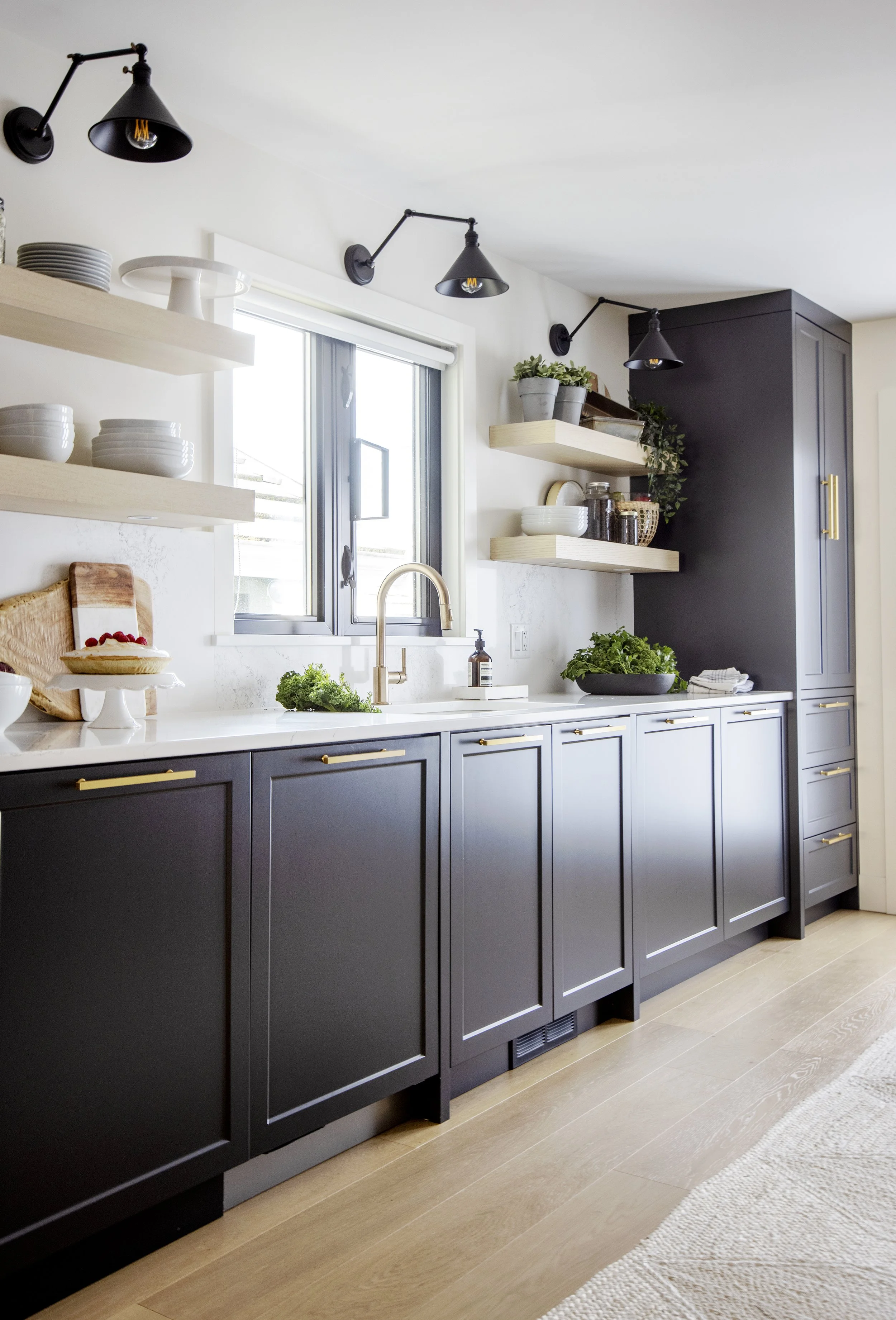 Modern kitchen with dark gray cabinets, white countertops, open shelves holding dishes, a window above the sink, and black adjustable wall lamps.