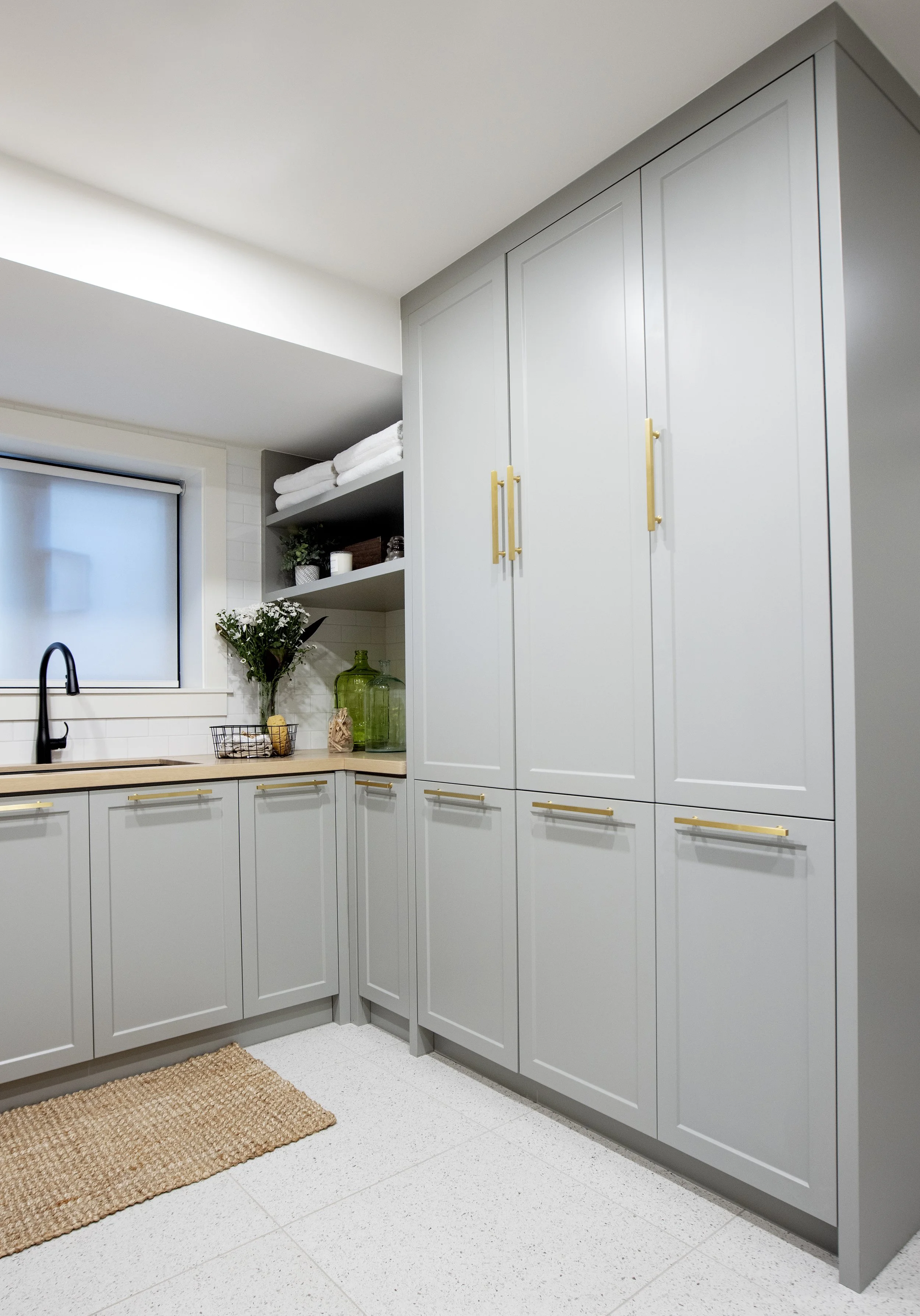 Kitchen with light gray cabinets, gold handles, white tiled backsplash, black faucet, open shelves with white towels, potted plants, and decorative glass bottles, beige rug, white speckled floor, and a window with a black frame.