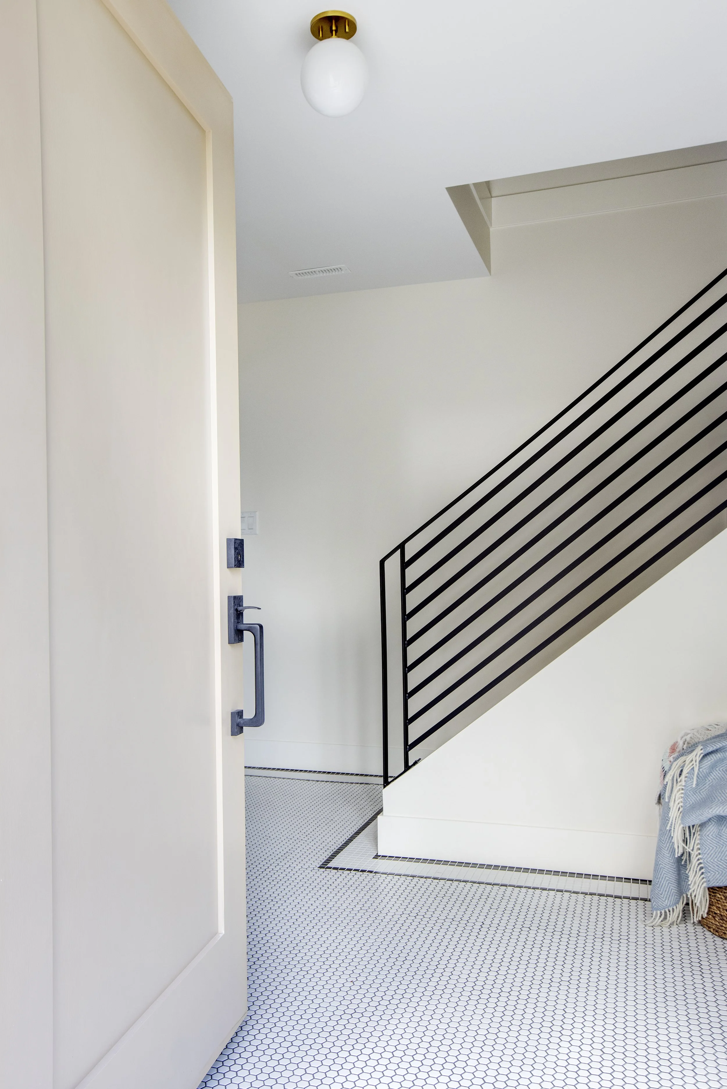 Interior view of a modern home with a white door, black metal staircase, and white hexagonal tiled flooring.